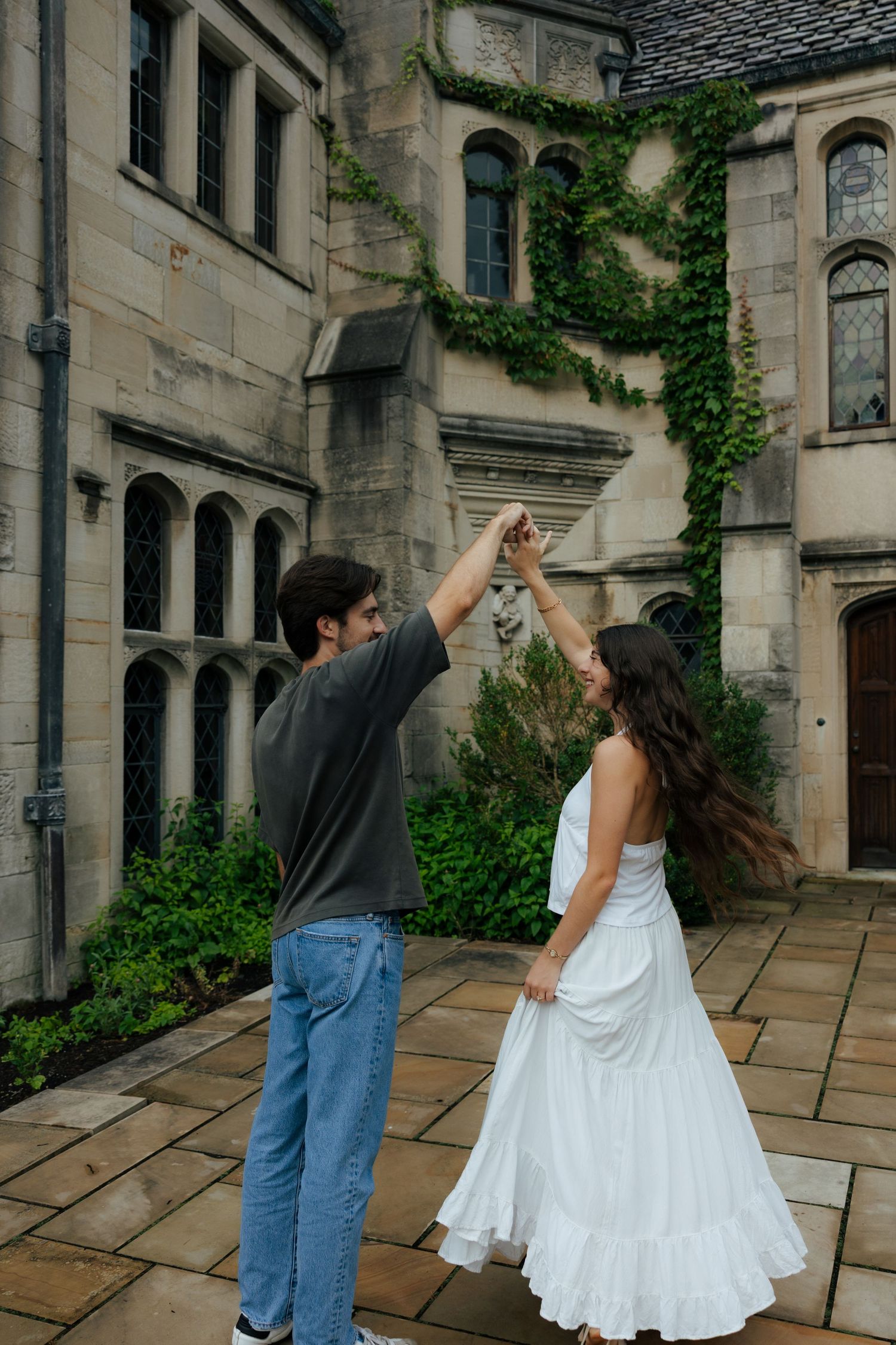 Man spinning woman in white dress outside historic stone building covered in ivy during engagement photos.