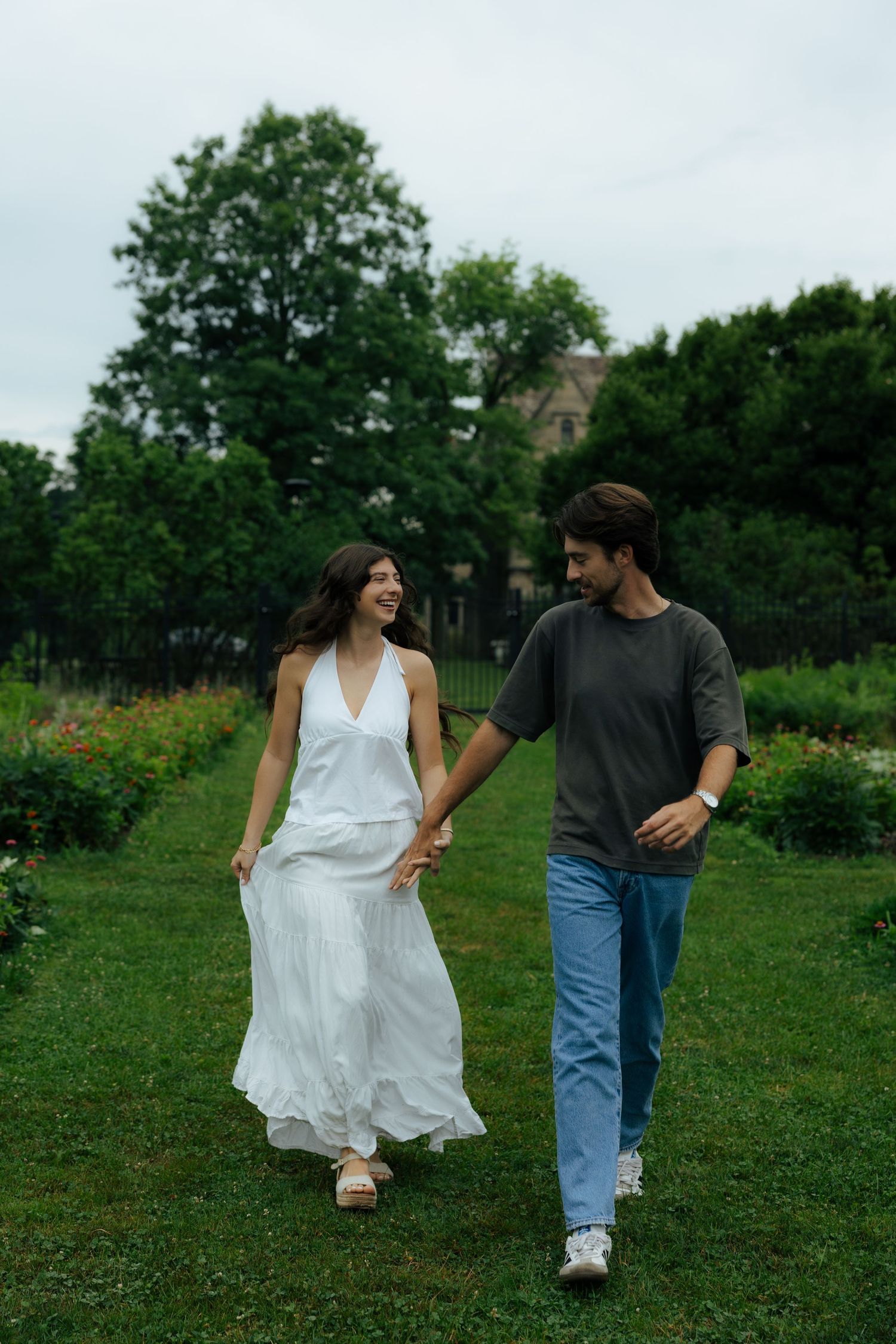 A smiling couple holds hands while walking through a lush green garden, woman in flowing white dress.