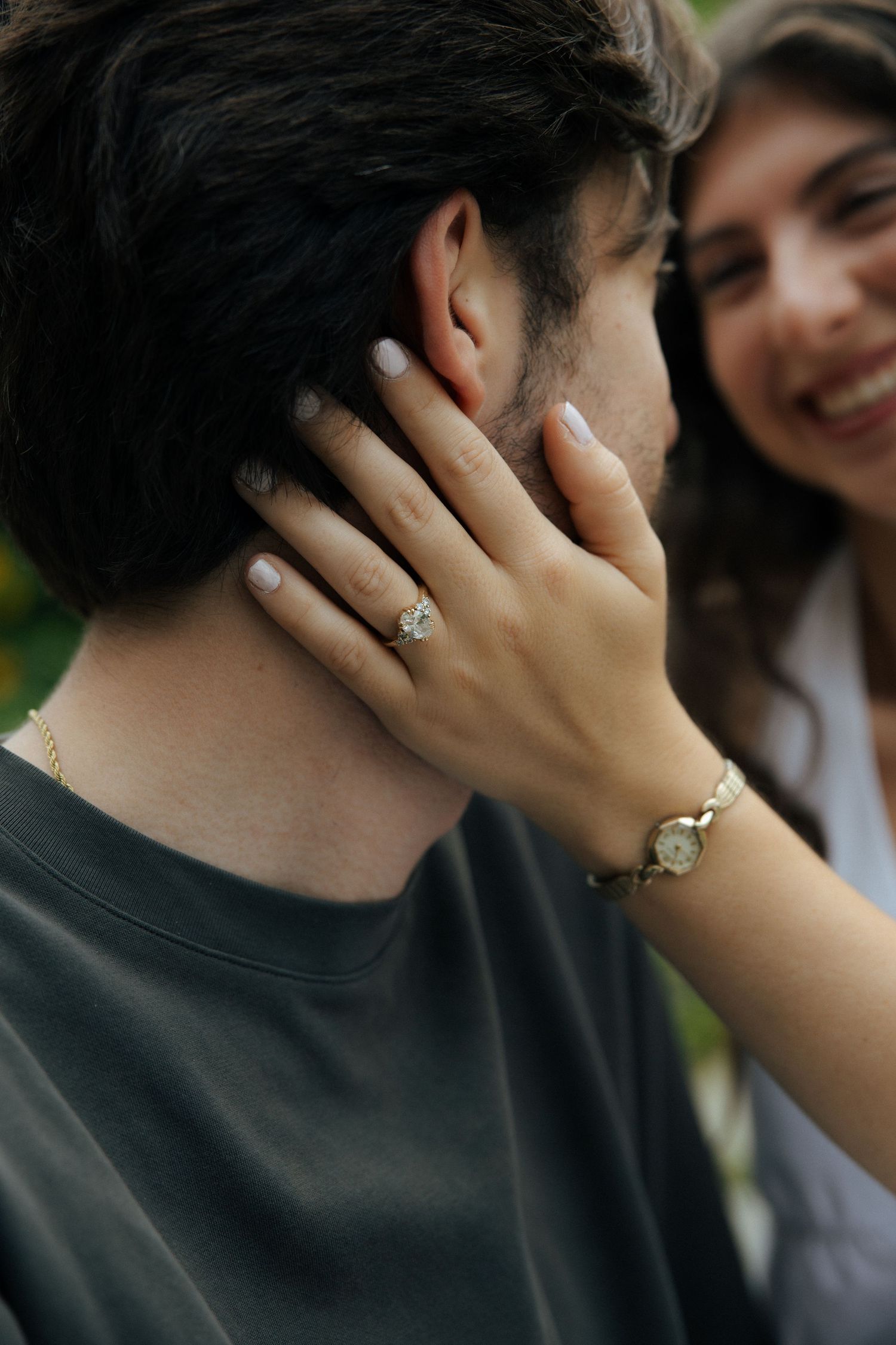 Woman with engagement ring gently touching man's face while smiling joyfully outdoors.