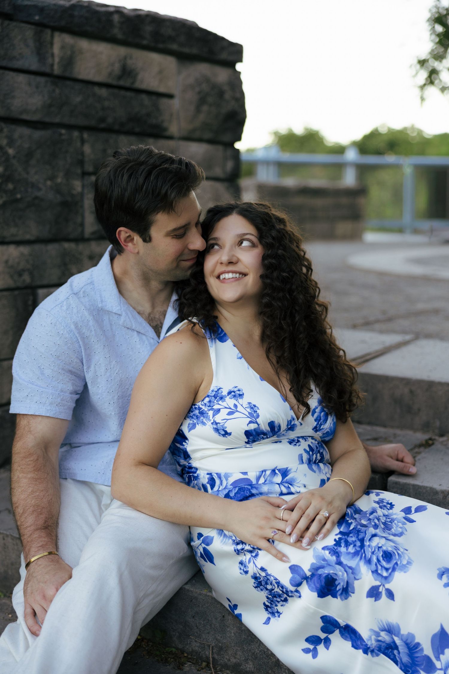 Happy couple sitting on stone steps, woman in blue floral dress smiling at her partner outdoors.