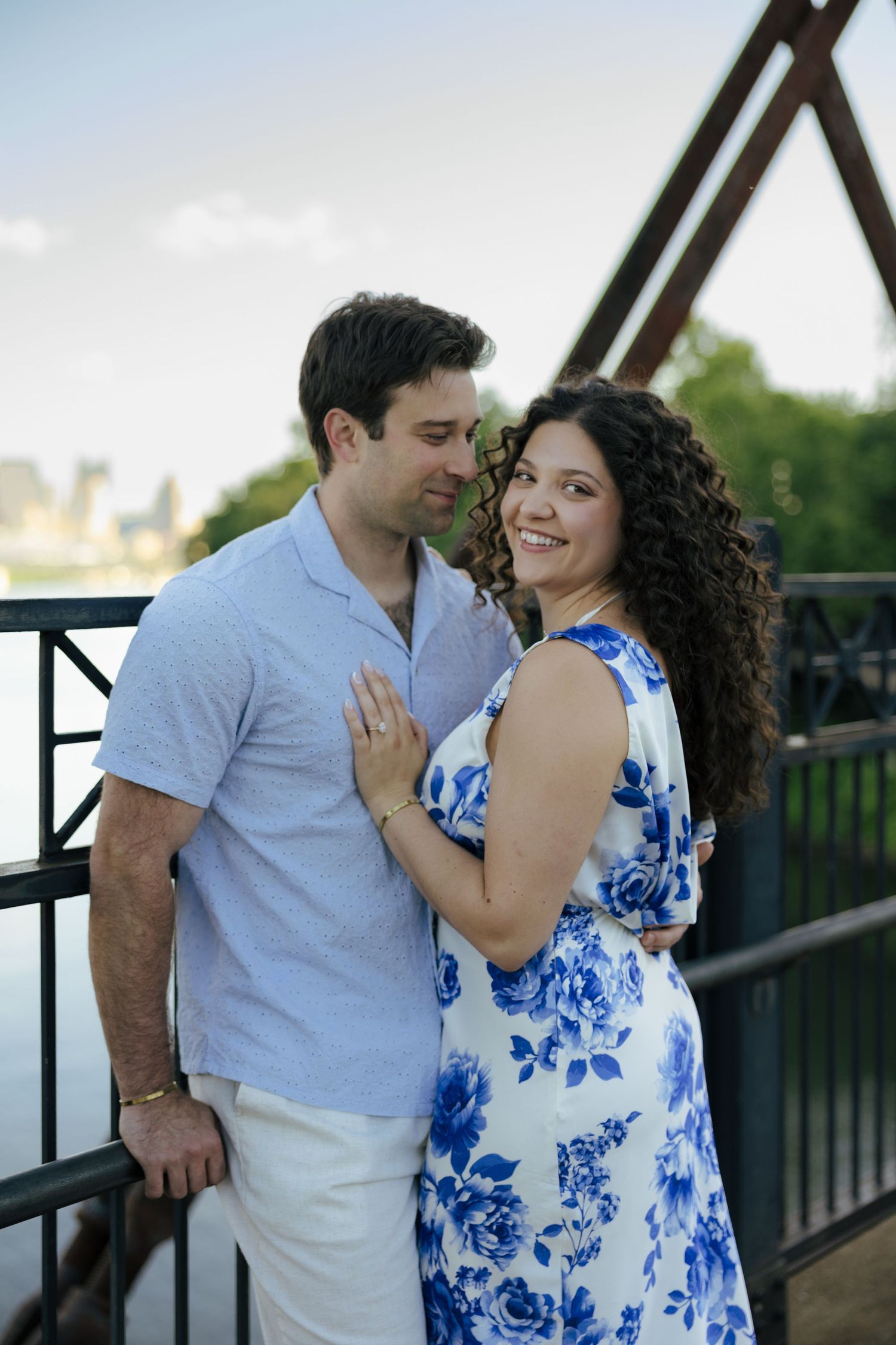 Couple embracing on a bridge, woman in blue floral dress smiling at camera, man in light blue shirt, city skyline behind.