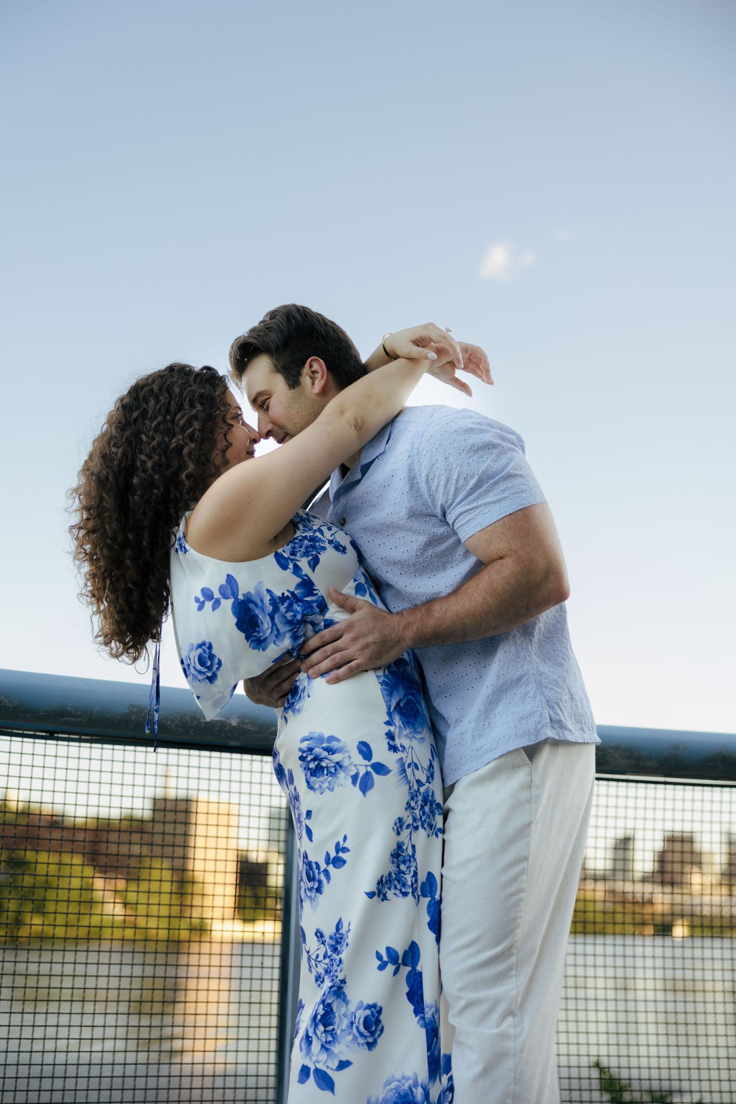 Romantic couple sharing a kiss on bridge railing, woman in blue floral dress at golden hour.