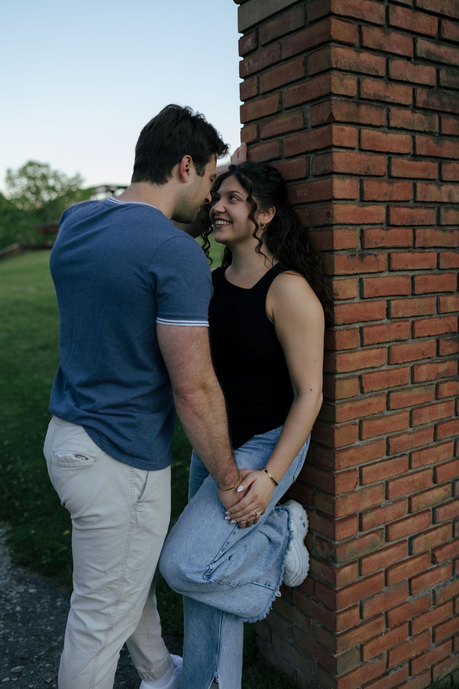A smiling couple holds hands and shares a romantic moment leaning against a red brick wall outdoors at dusk.