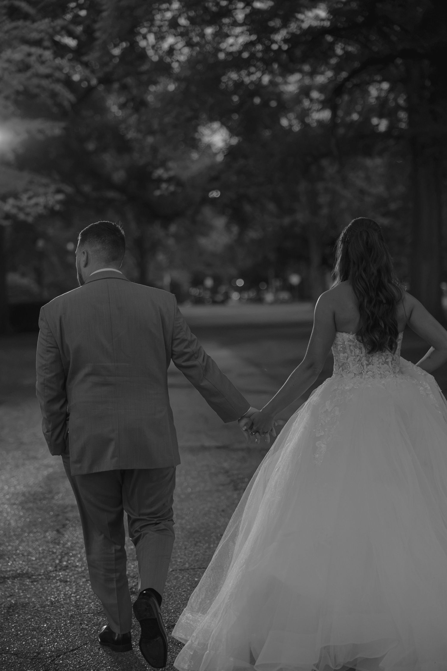 Black and white photo of a bride and groom holding hands walking down a tree-lined path on their wedding day.