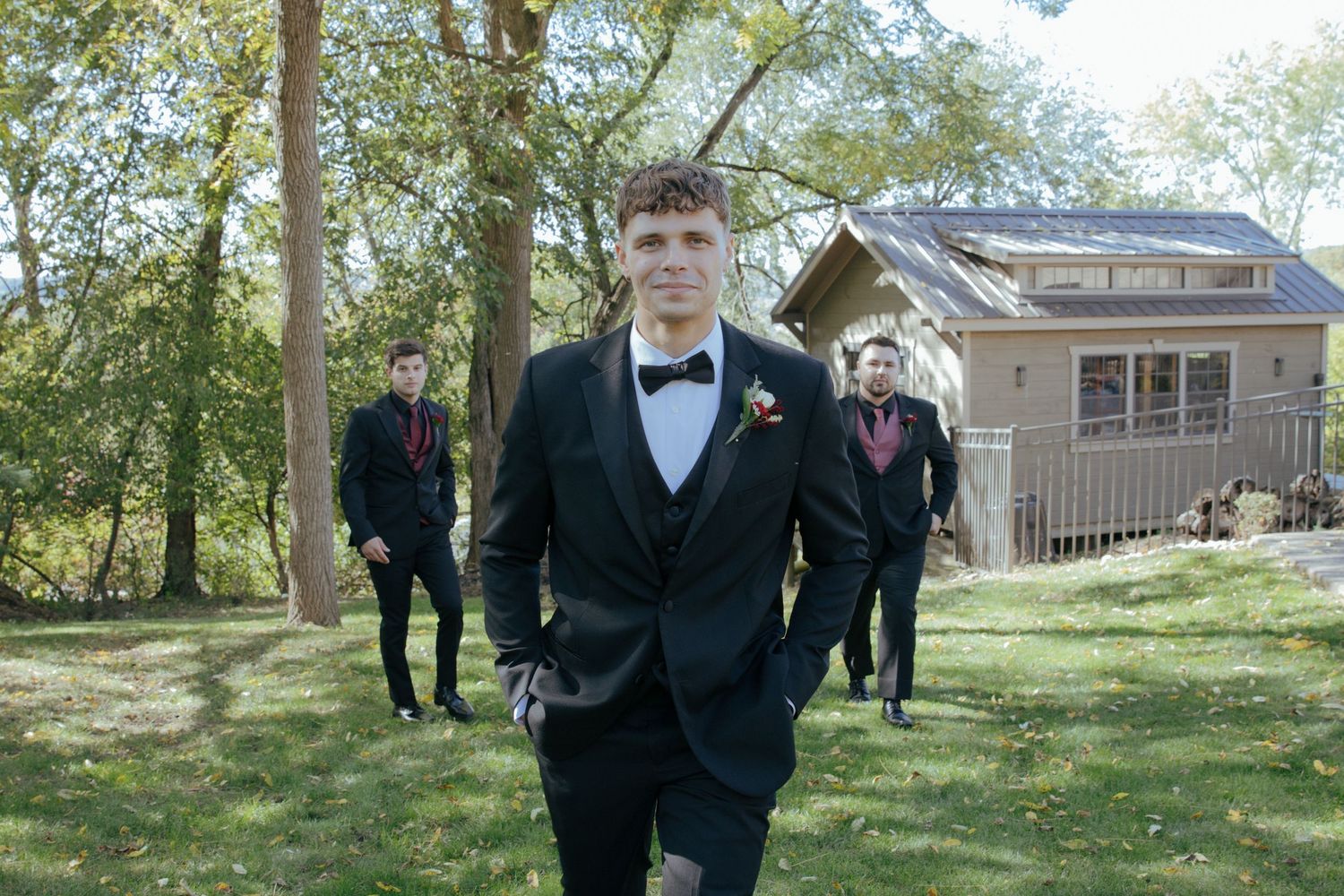 Young men in black tuxedos with red boutonnieres walk outdoors near a house surrounded by trees.