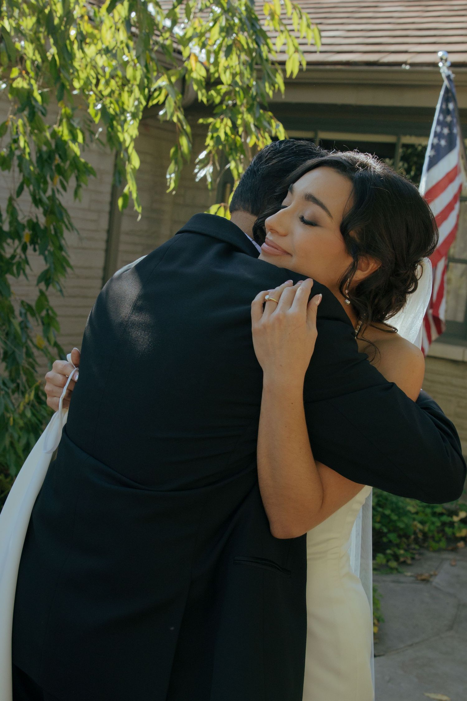 Bride and groom share a tender embrace outdoors, with an American flag visible in the background.