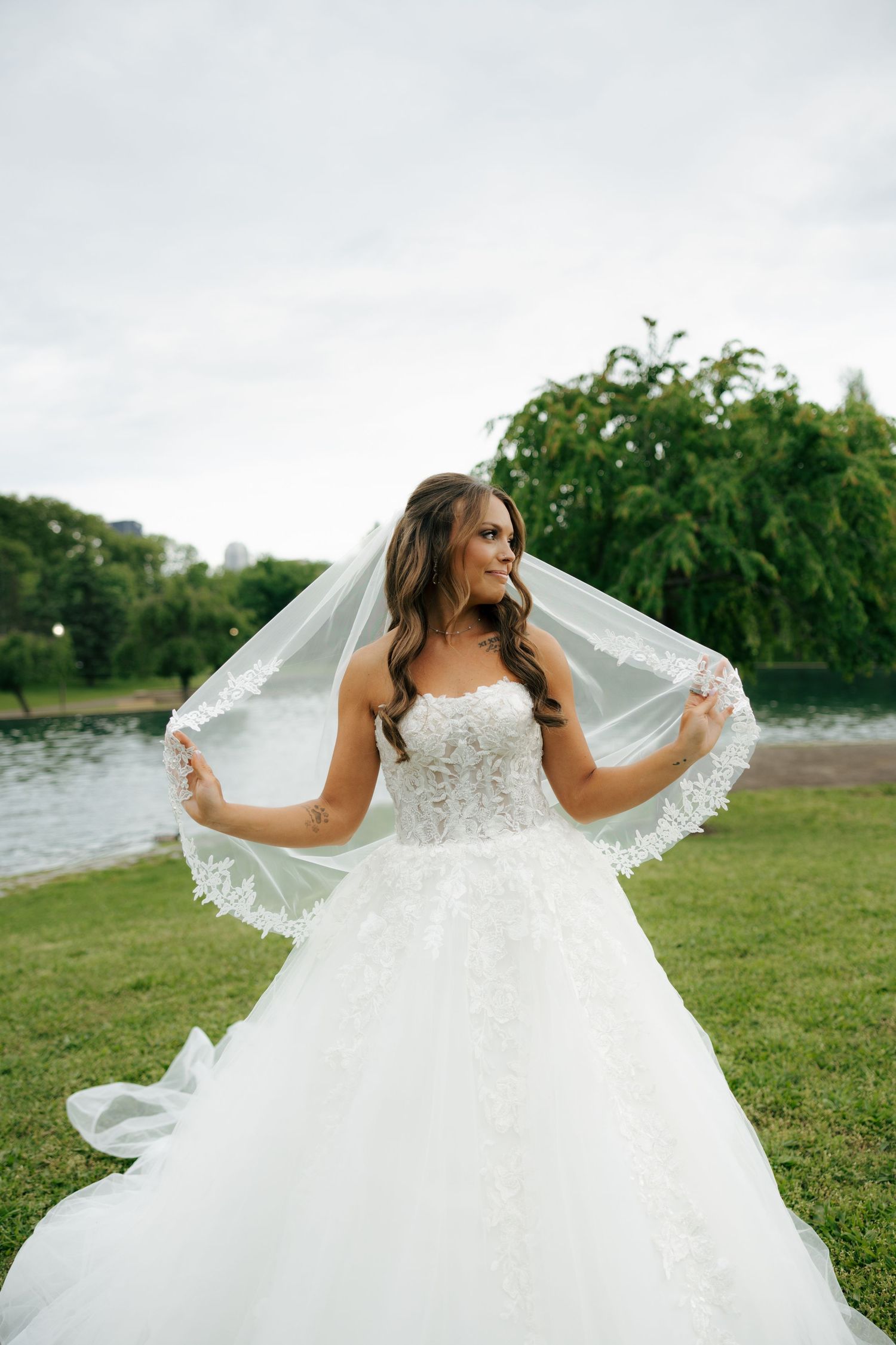 Bride in a strapless lace ballgown holding her veil up outdoors by a lake with lush green trees.