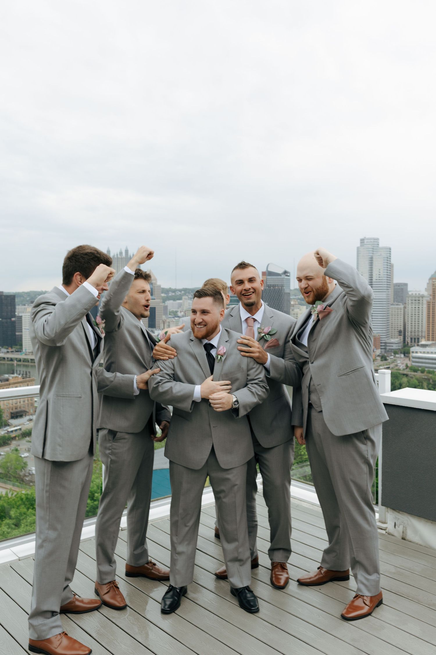 Groom and groomsmen in gray suits celebrate on a rooftop with a city skyline backdrop.
