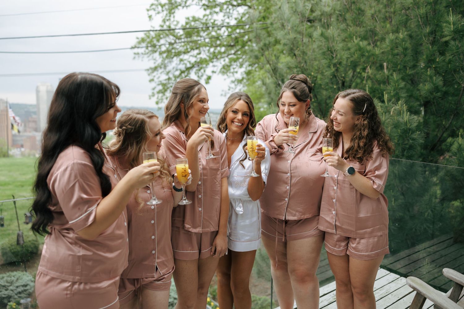 Bride in white satin pajamas celebrates with bridesmaids in matching pink sets, holding champagne glasses on an outdoor deck.