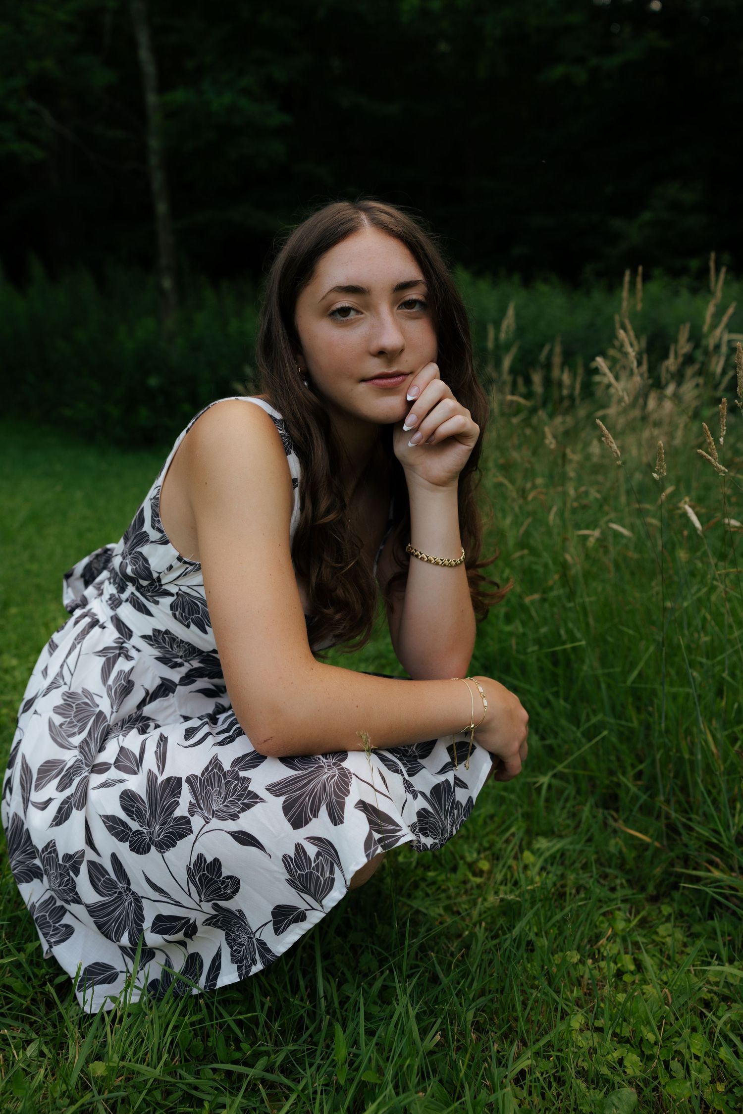 Young woman in white floral dress posing thoughtfully in tall grass with dark forest background.