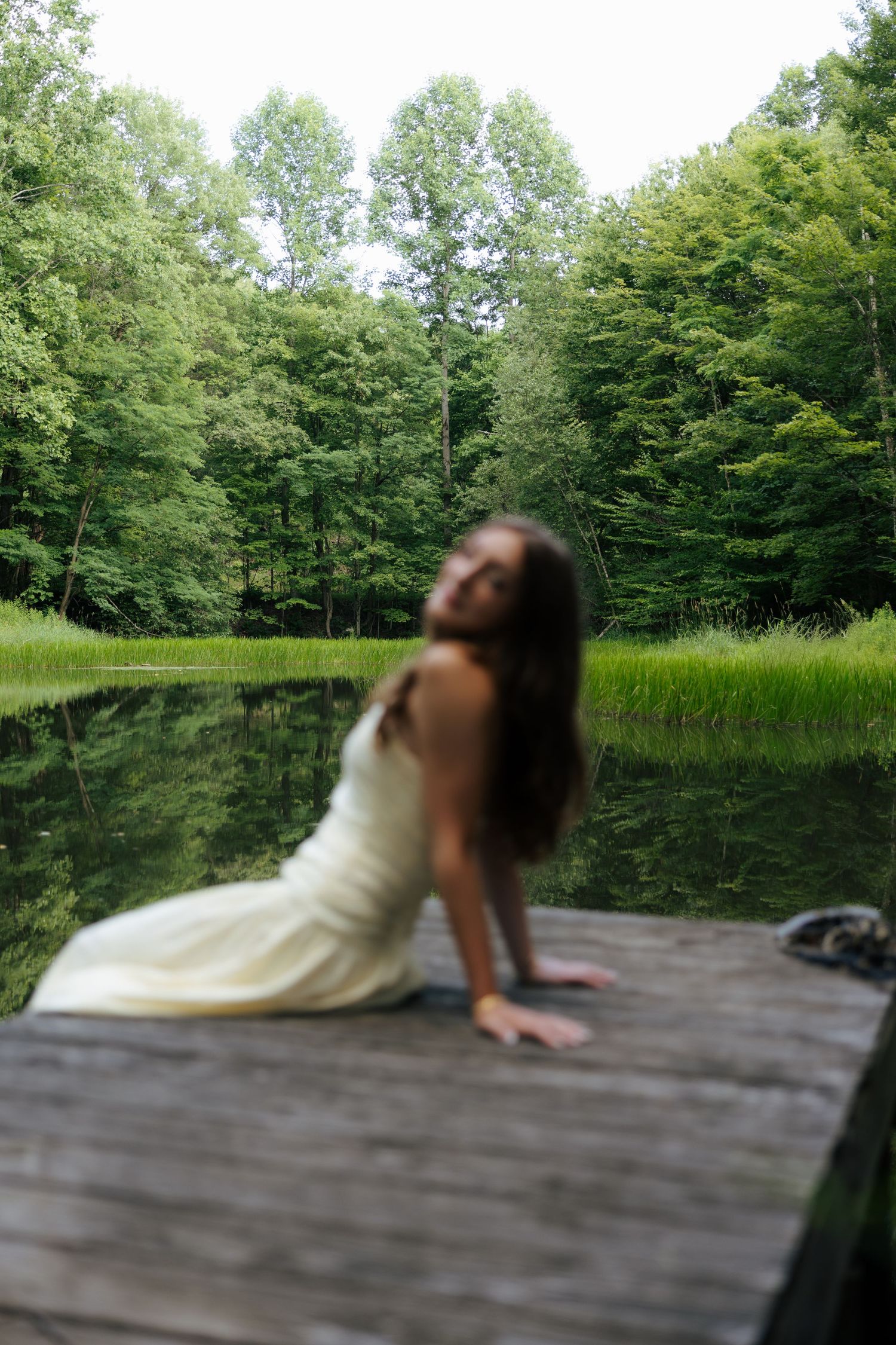 Young woman in white dress posed on rustic dock with blurred green forest and lake background.