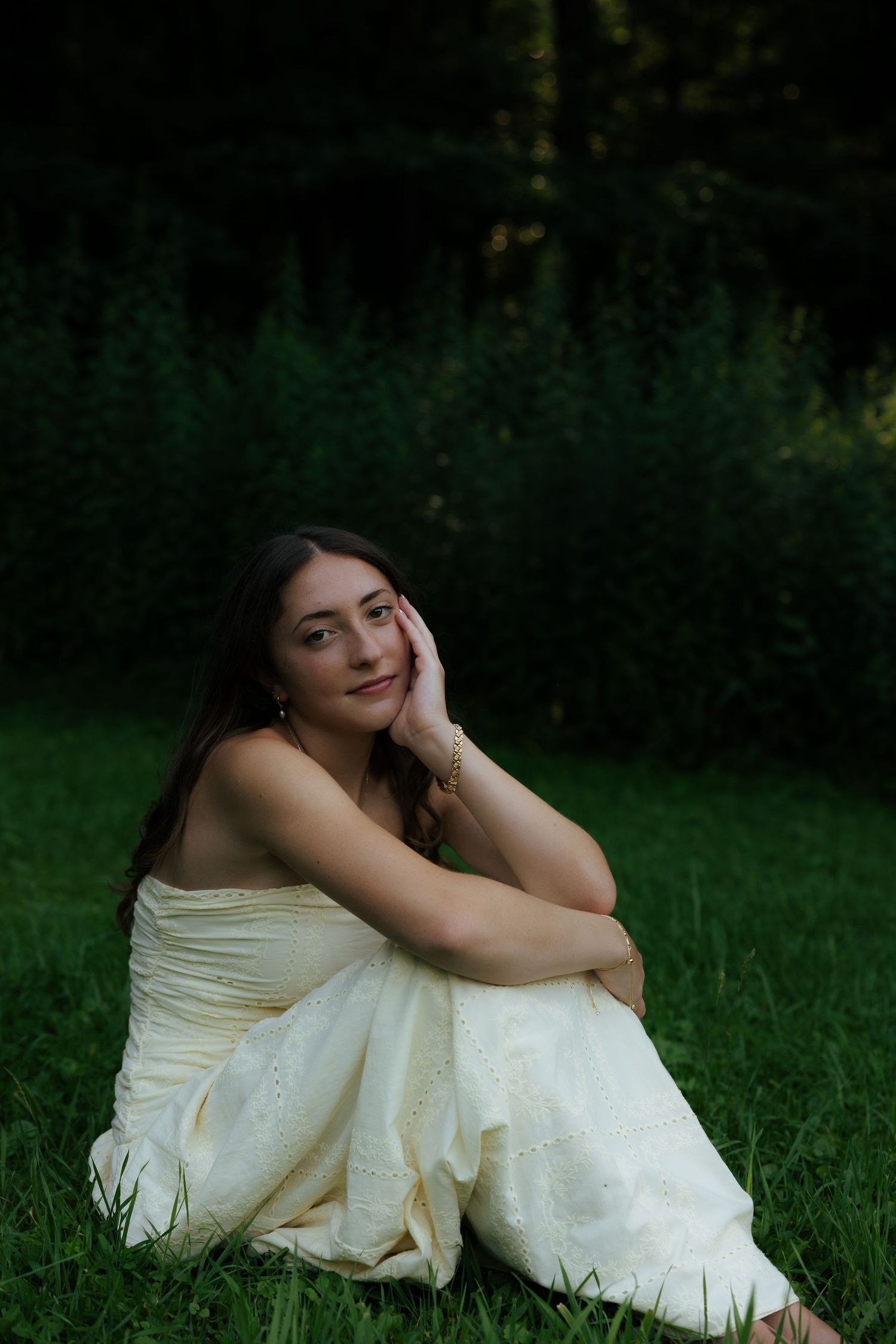 Young woman in cream dress sitting on grass, smiling softly with hand resting on cheek outdoors.