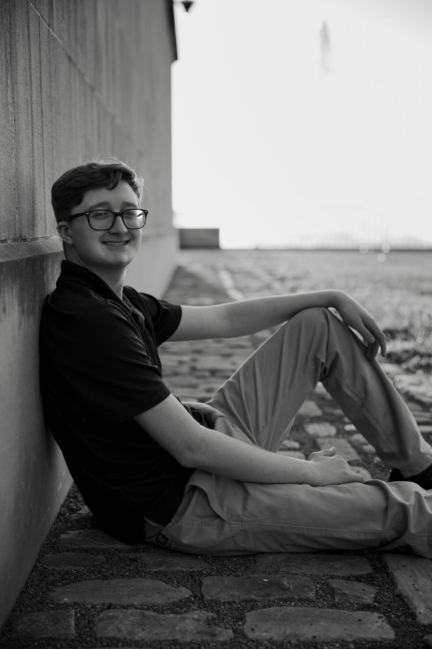 Black and white portrait of smiling teen in glasses sitting against wall on cobblestone ground.