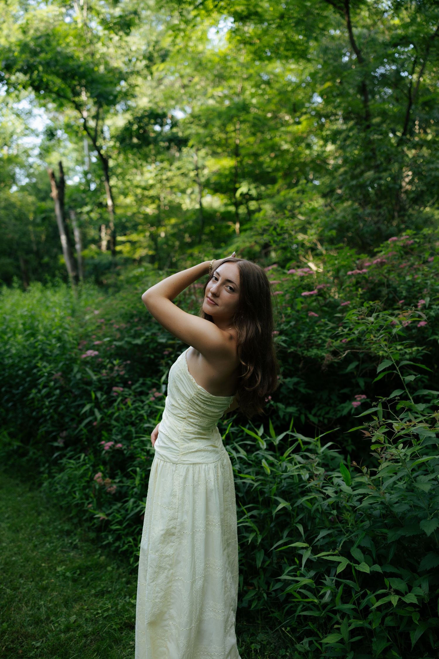 Young woman in elegant white maxi dress posing among green foliage in a lush forest setting.