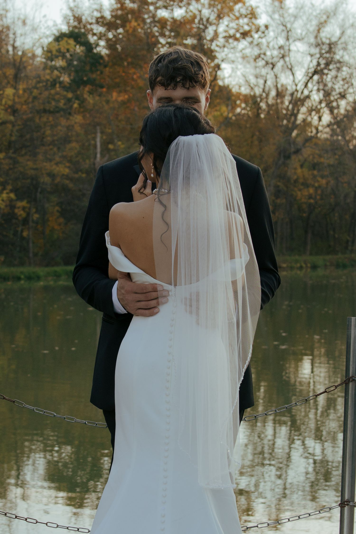 Bride in backless gown and veil embraces groom near a serene autumn lake during wedding photo session.