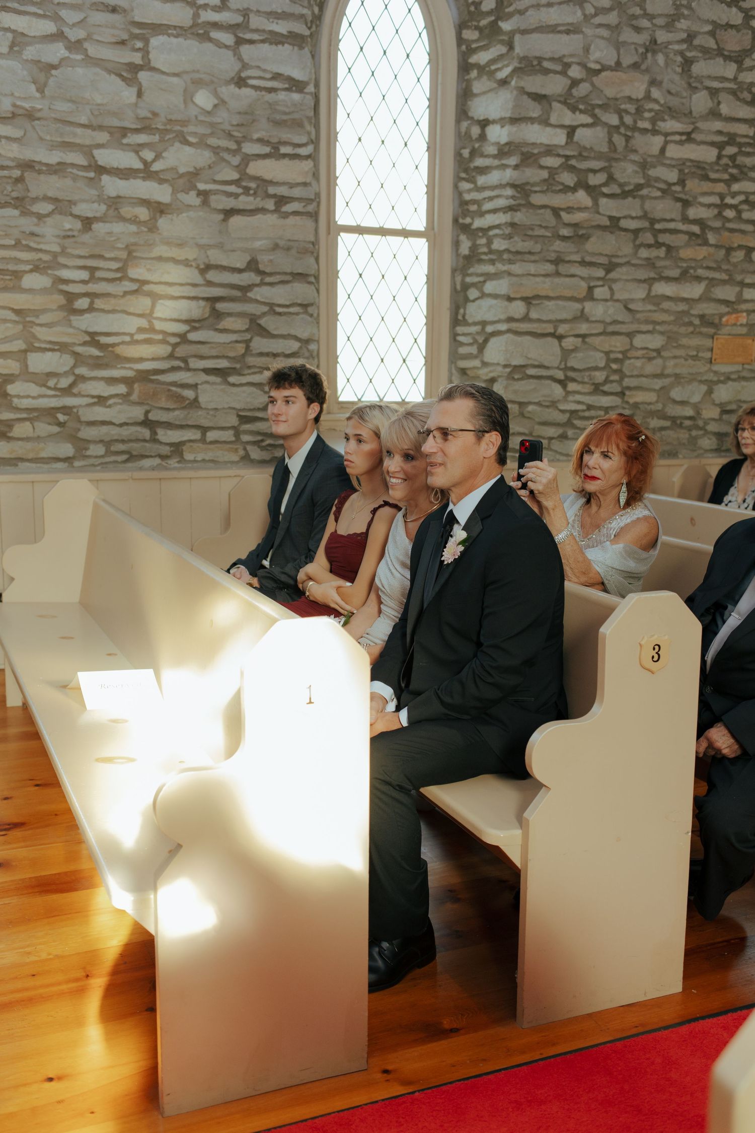 Wedding guests seated in white wooden pews inside a rustic stone chapel during the ceremony.