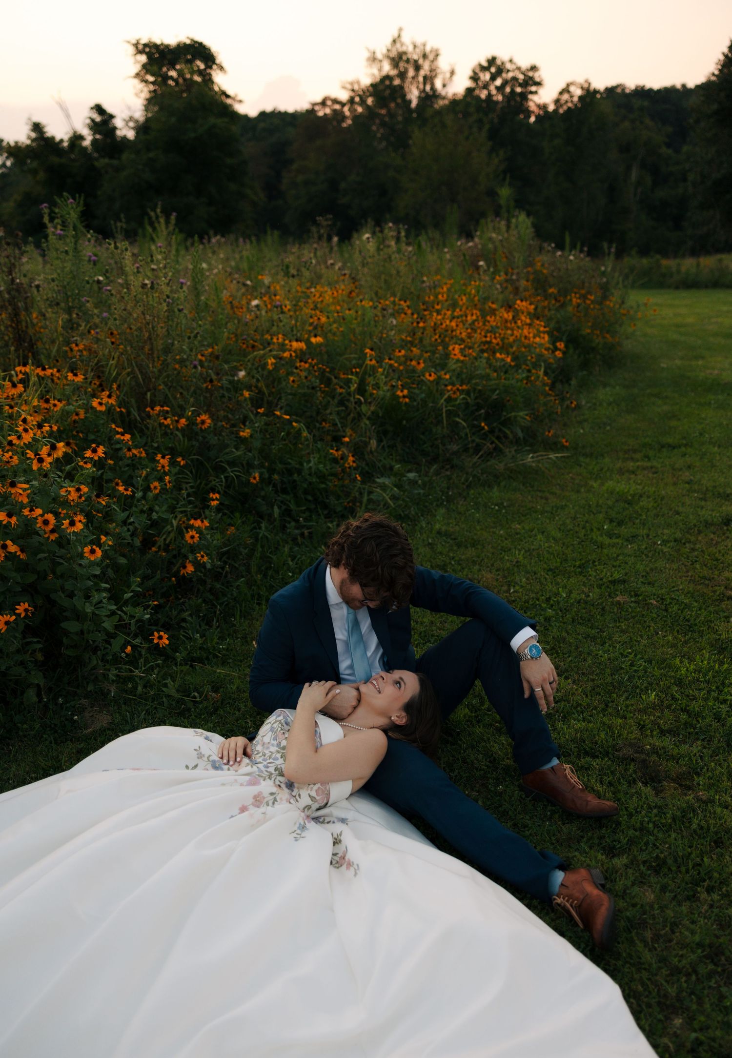 Romantic couple poses at dusk, bride reclines on grass near vibrant orange wildflowers as groom leans in.