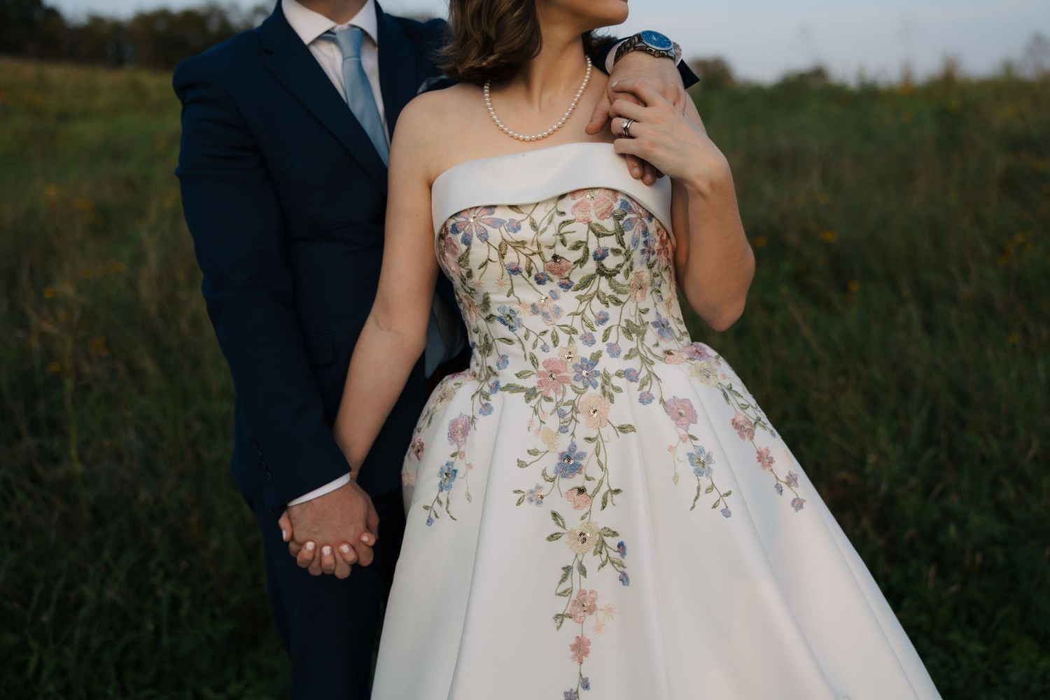 Bride in floral embroidered strapless ballgown holds hands with groom in navy suit in a green field.
