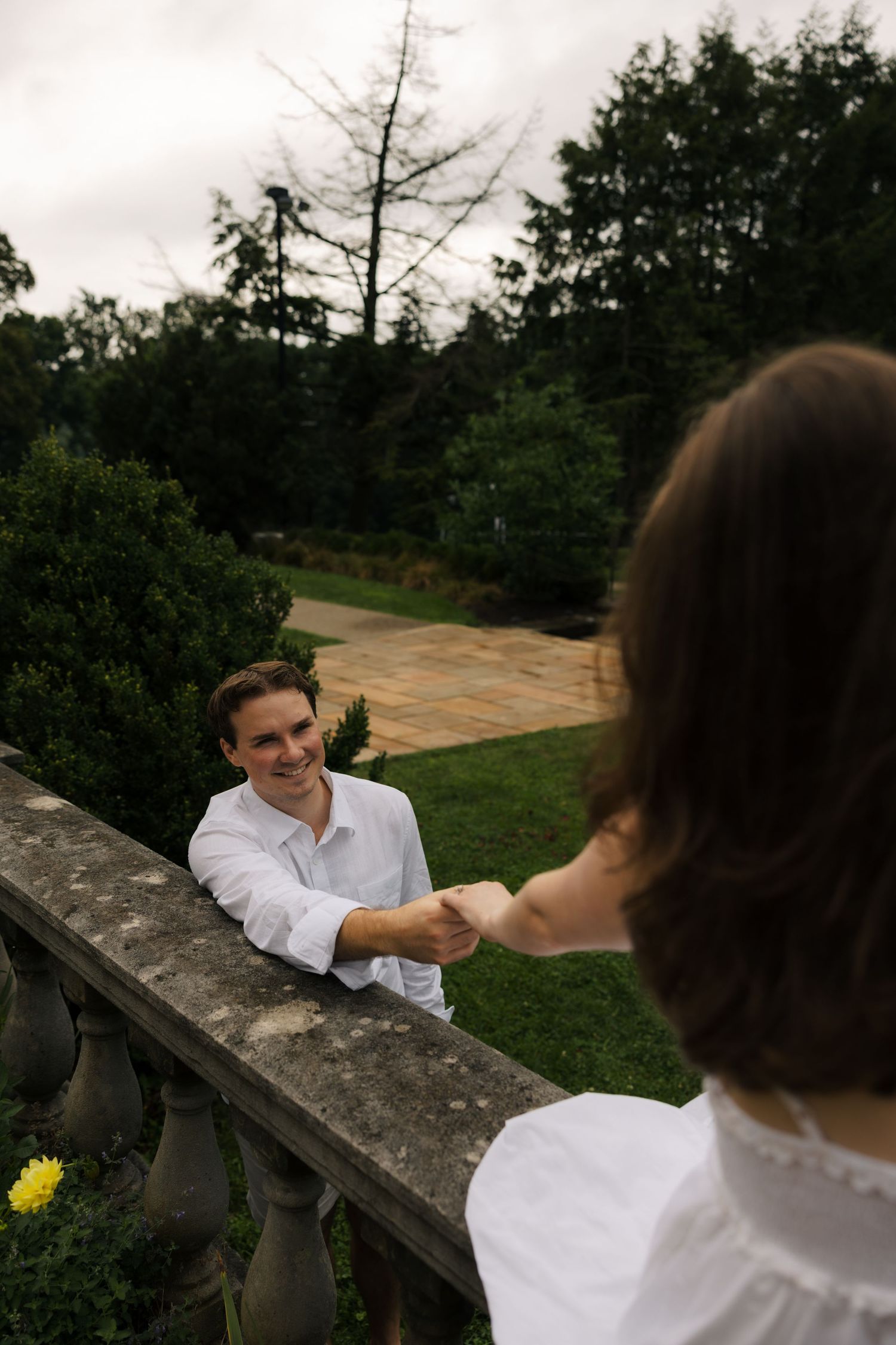 Smiling man in white shirt holds woman's hand over stone balustrade in lush garden setting.