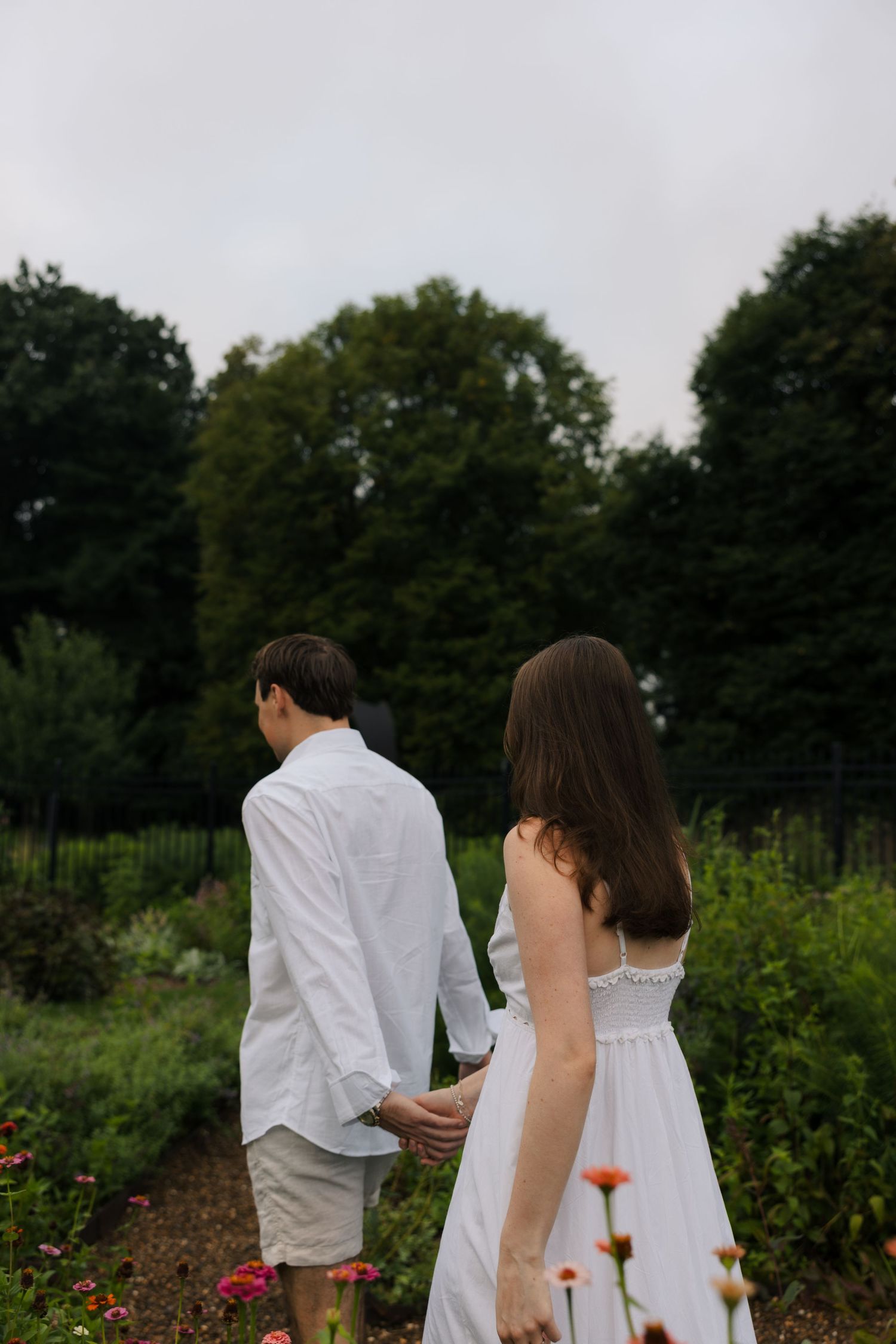 A couple in white outfits holds hands while walking through a lush garden with colorful flowers and tall trees.