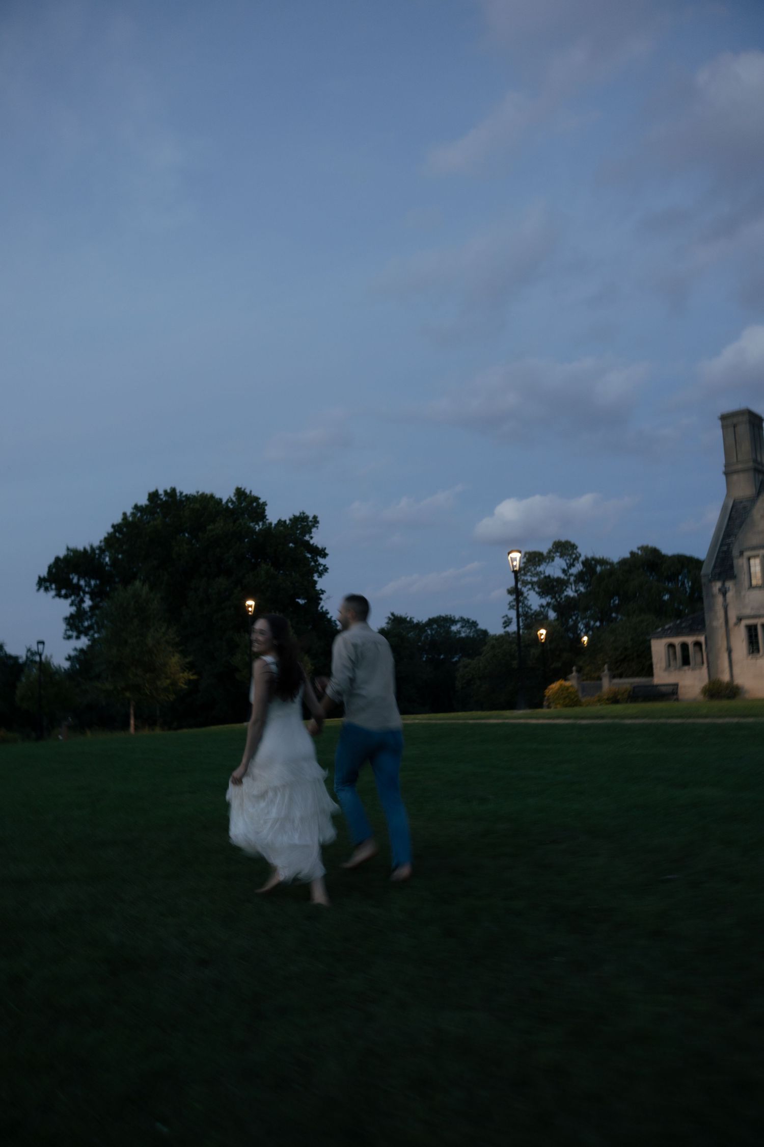 Couple holding hands walking across a green lawn at twilight near a lit historic building under a blue sky.