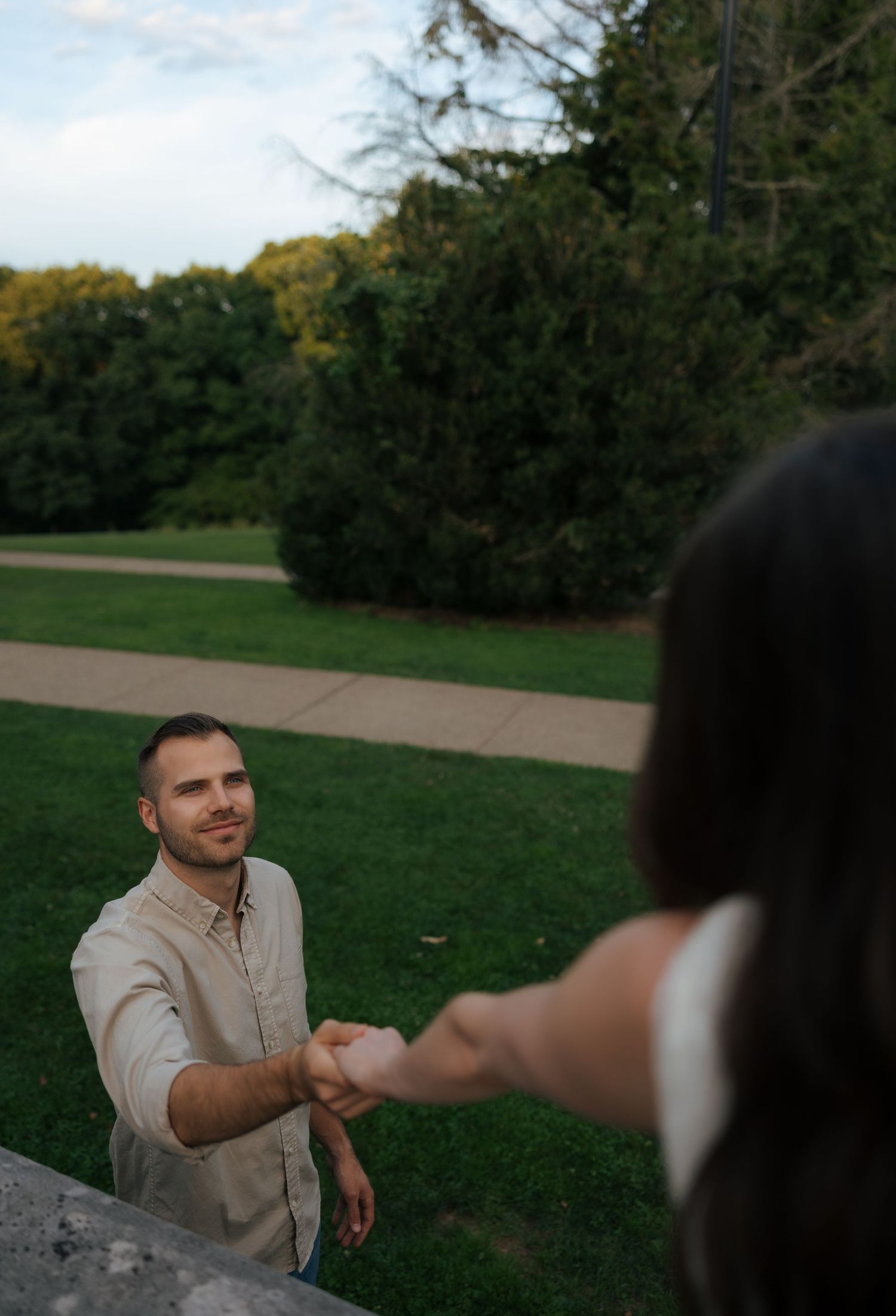 Smiling man in beige shirt reaches out to hold hands with woman during outdoor engagement photo session.