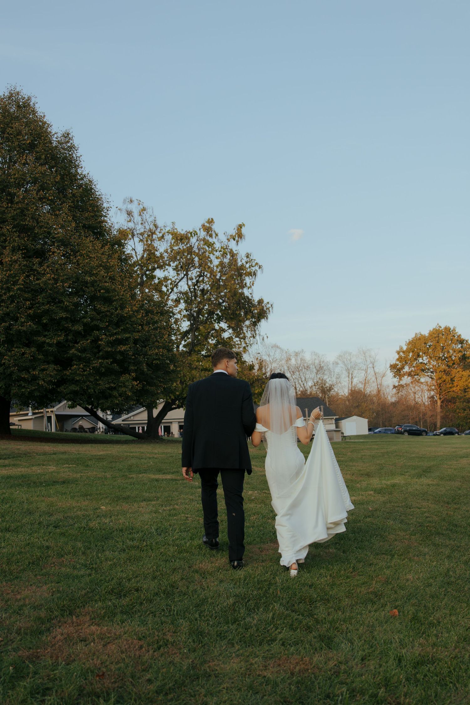 Bride and groom walking hand in hand across a green lawn at sunset with trees in the background.