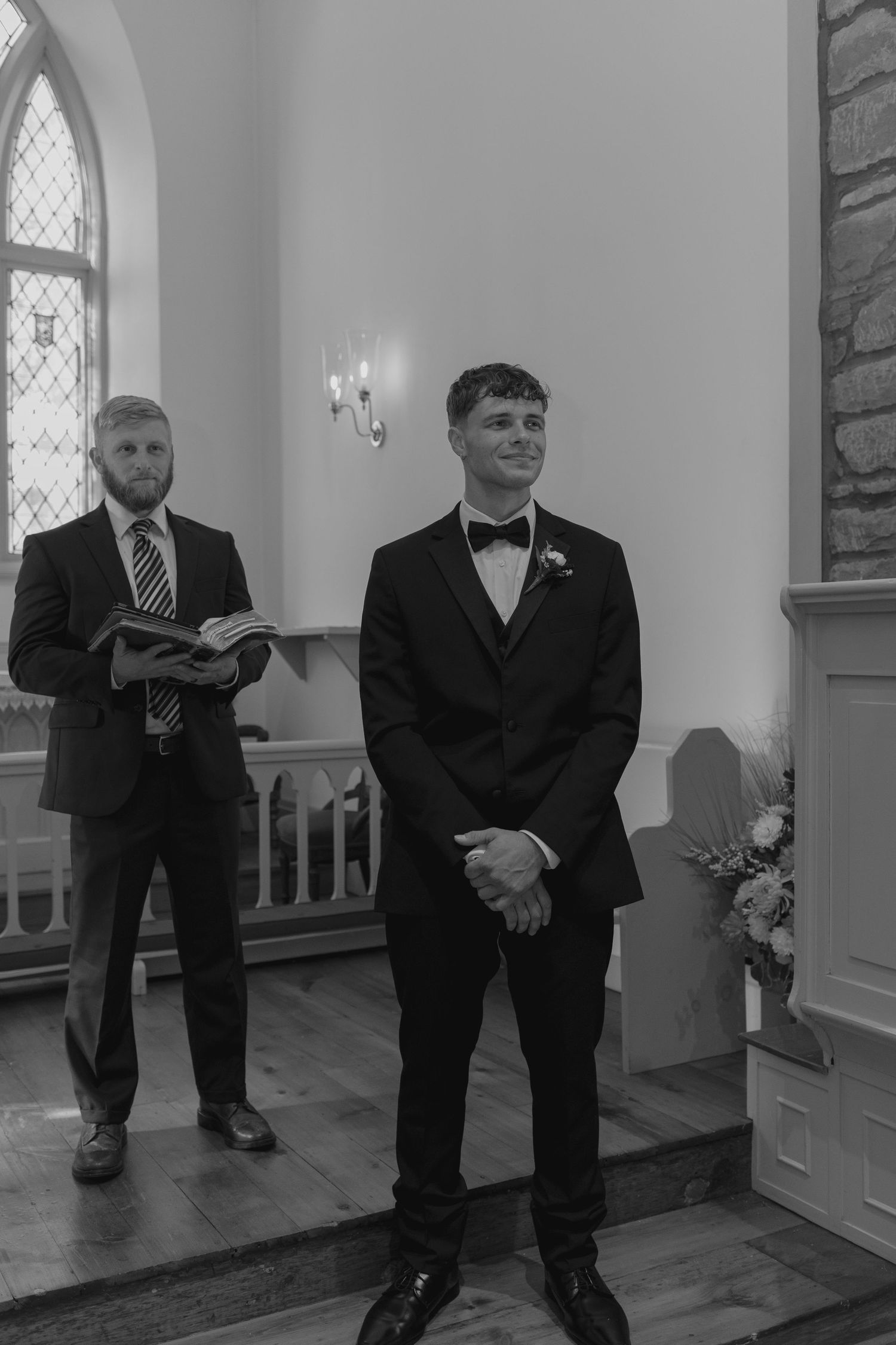 Groom in black tuxedo with bow tie smiles at altar as officiant holds open book during wedding ceremony.