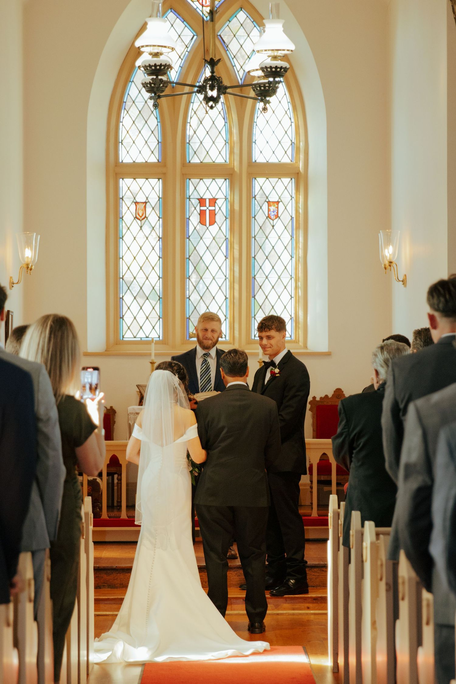 Bride and groom exchange vows at church altar with stained glass windows and wedding guests seated in pews.