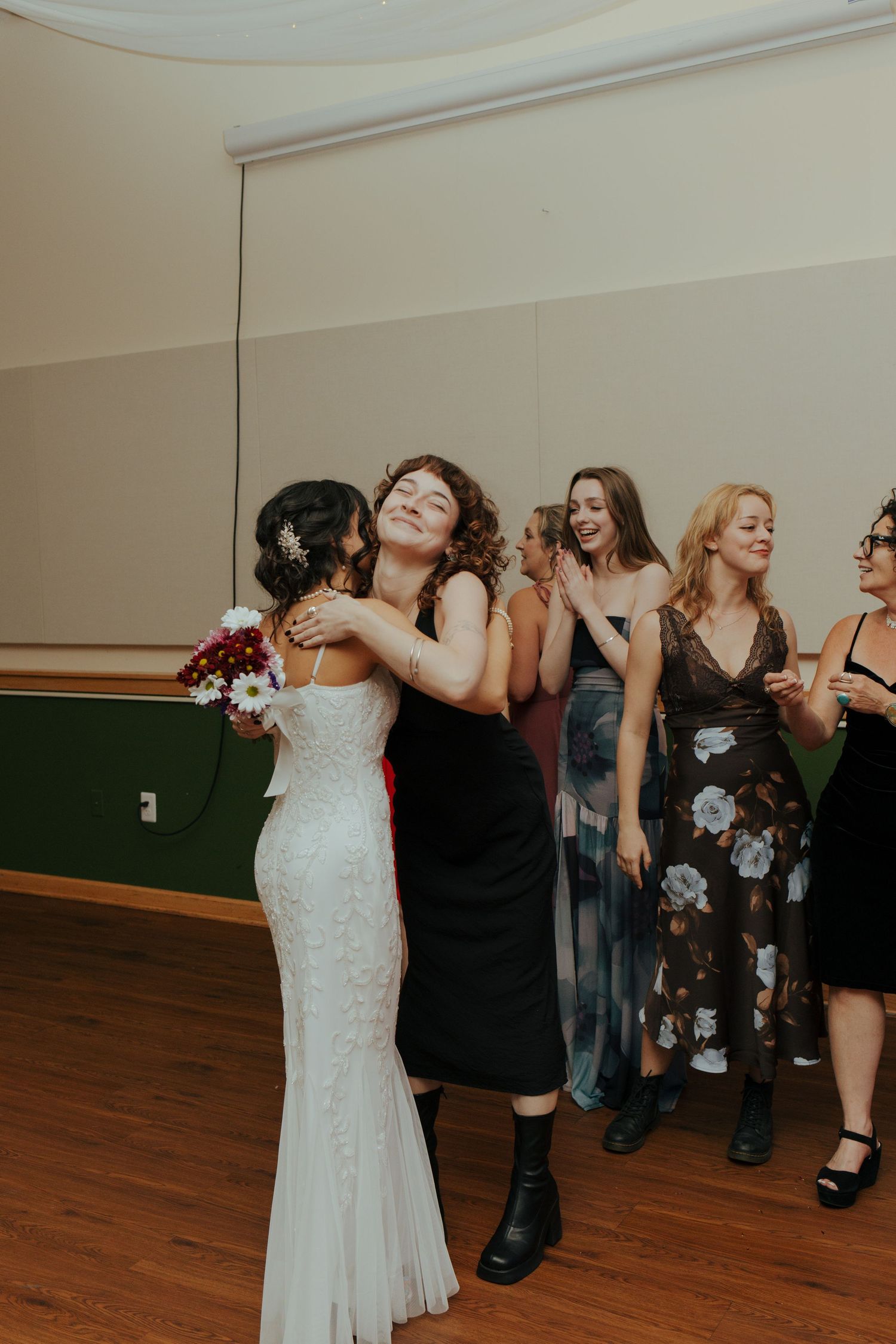 Bride in white gown hugging curly-haired guest while bridesmaids laugh during bouquet toss at reception.