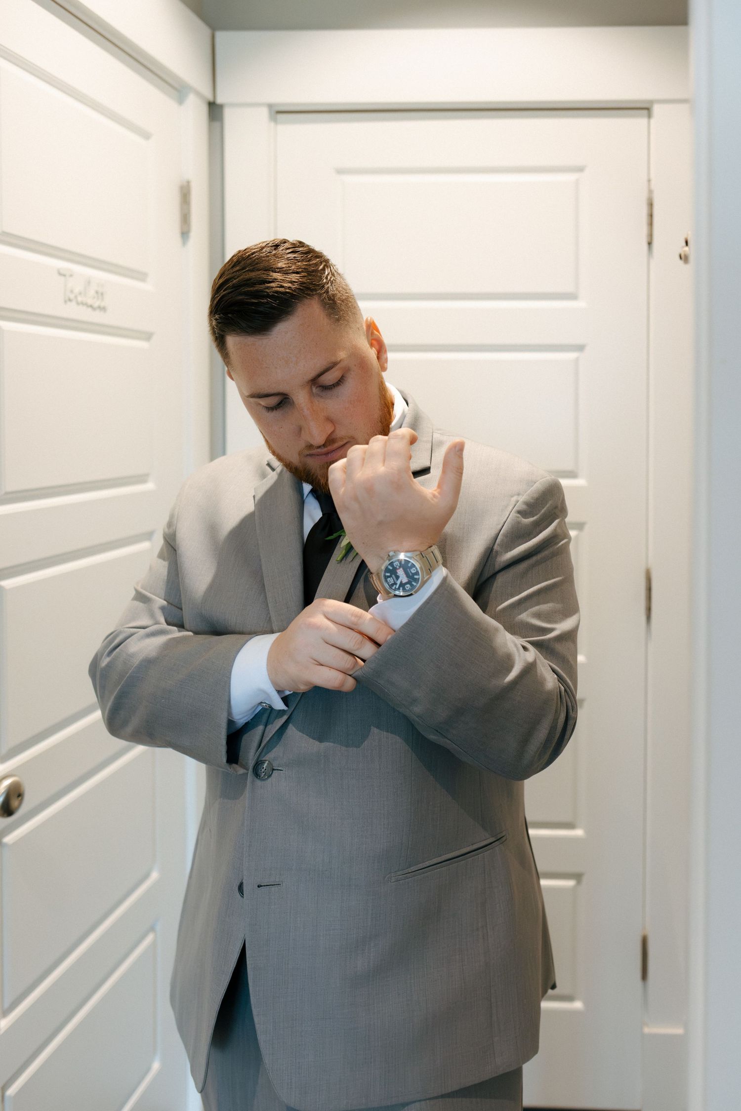 A well-dressed groom in a gray suit adjusts his cufflinks while wearing a blue watch, preparing for his wedding day.