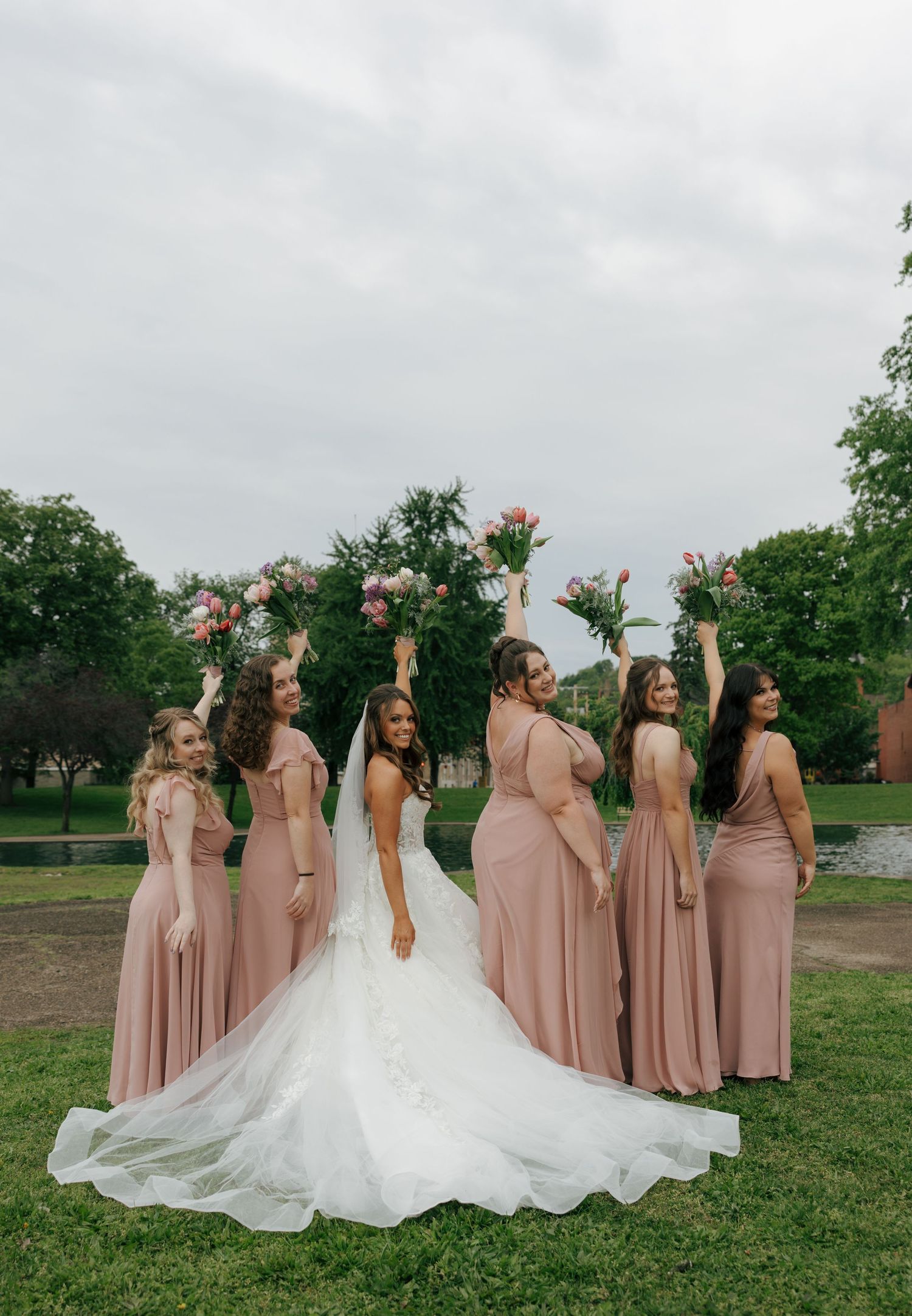 Bride in white gown poses with bridesmaids in blush dresses raising floral bouquets.