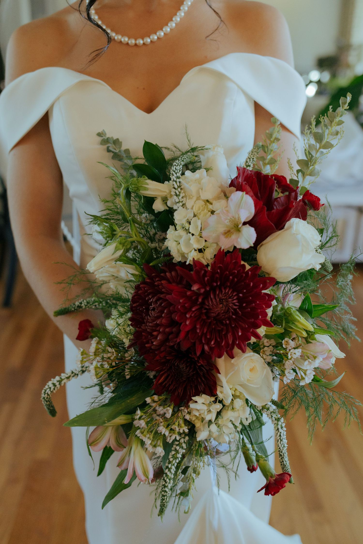 Bride holding lush cascading bouquet of dark red dahlias and white flowers in off-shoulder gown.