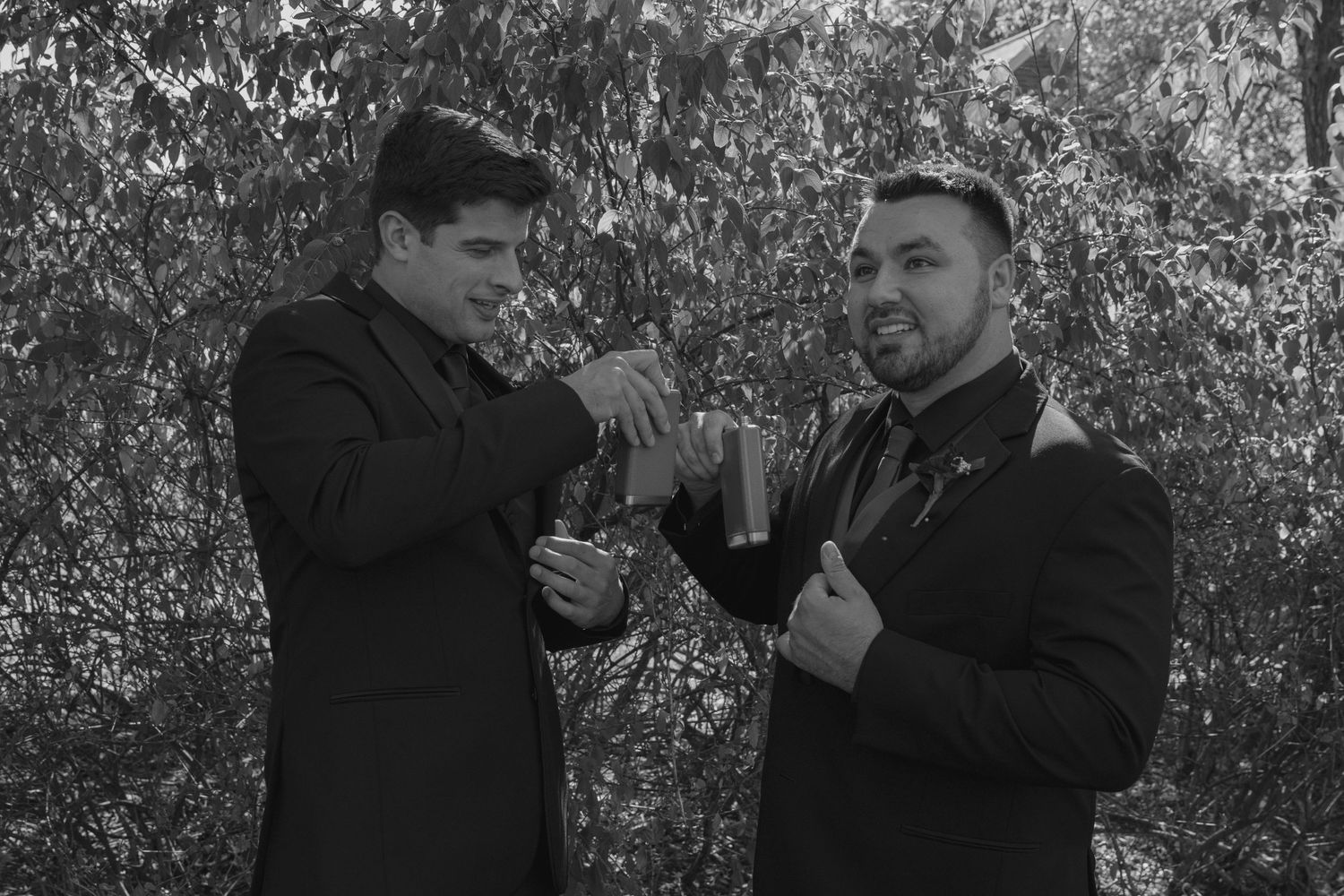 Two men in black suits sharing a toast outdoors in a black and white wedding photo.