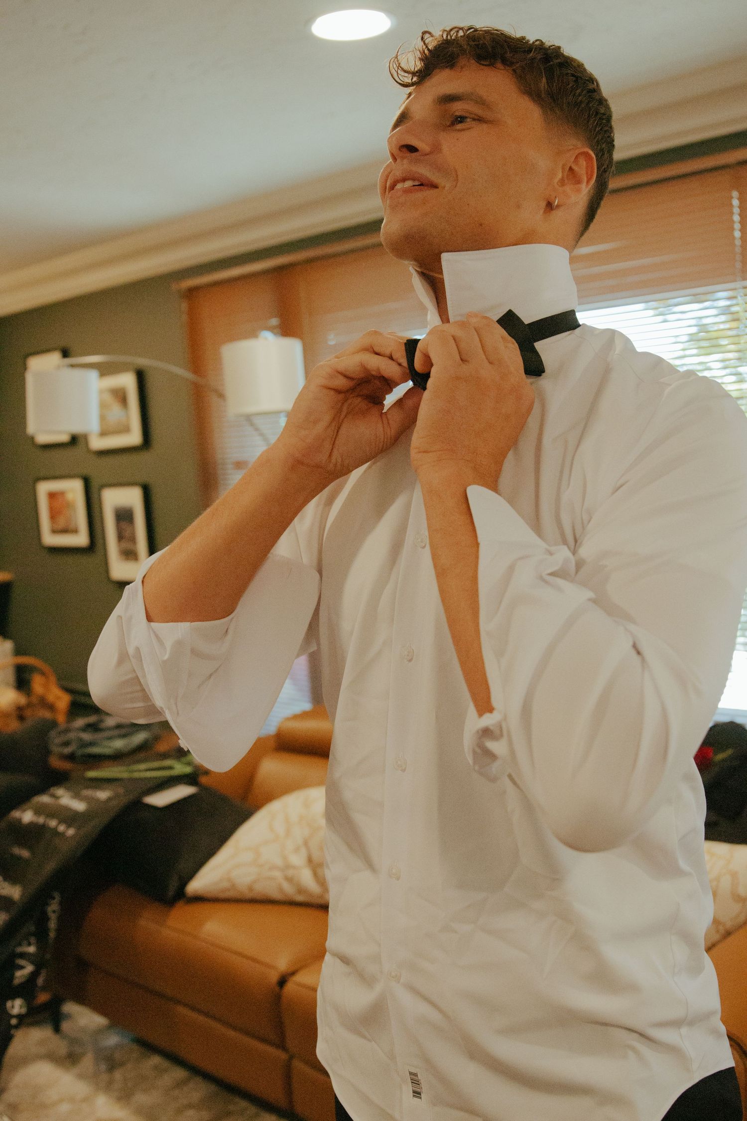 Groom in white dress shirt smiling while adjusting black bow tie during wedding day preparations.