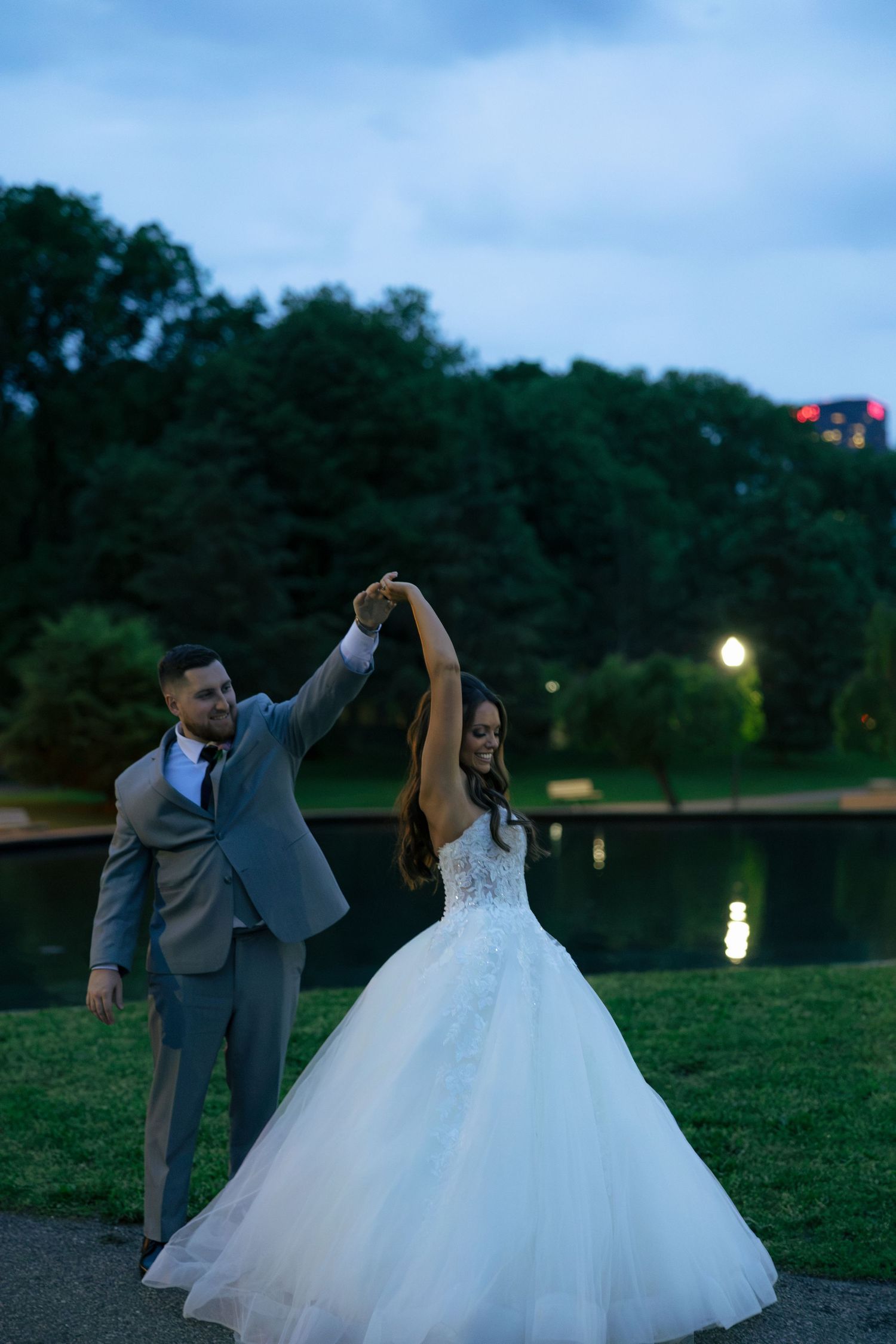 Bride and groom dancing outdoors at dusk near a pond, bride in a white ballgown, groom in a gray suit.