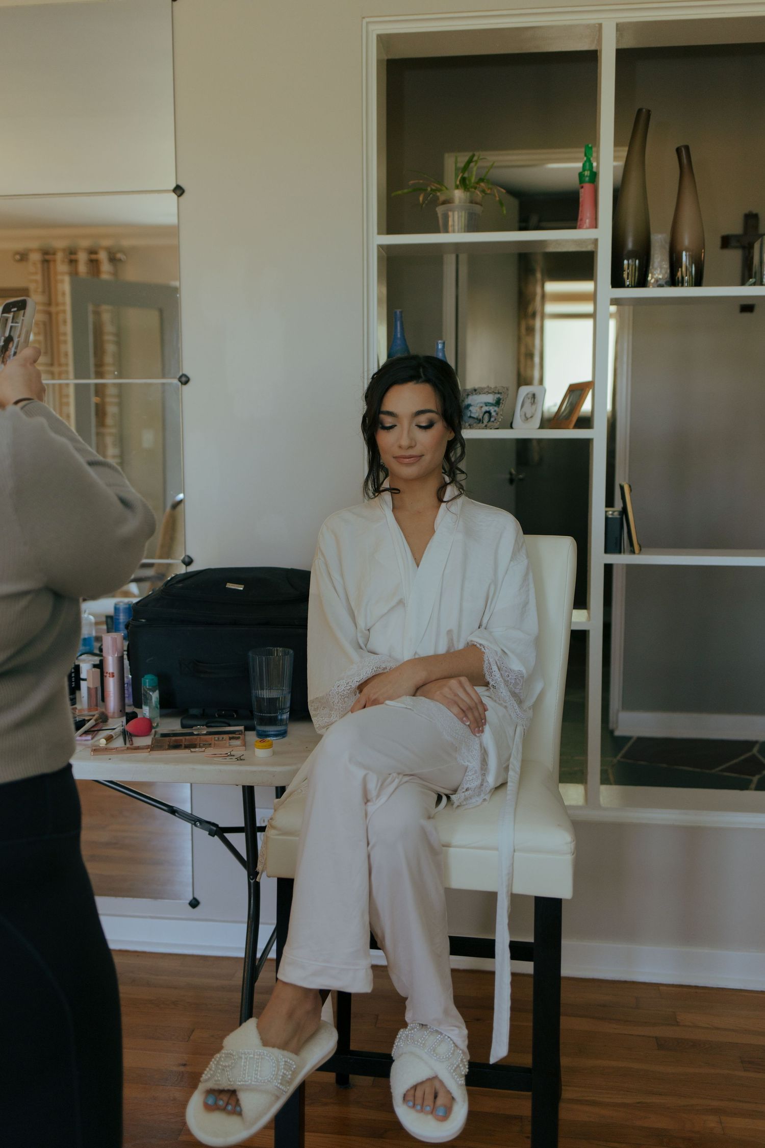 Bride in white robe and slippers sits in chair during wedding hair and makeup preparations indoors.