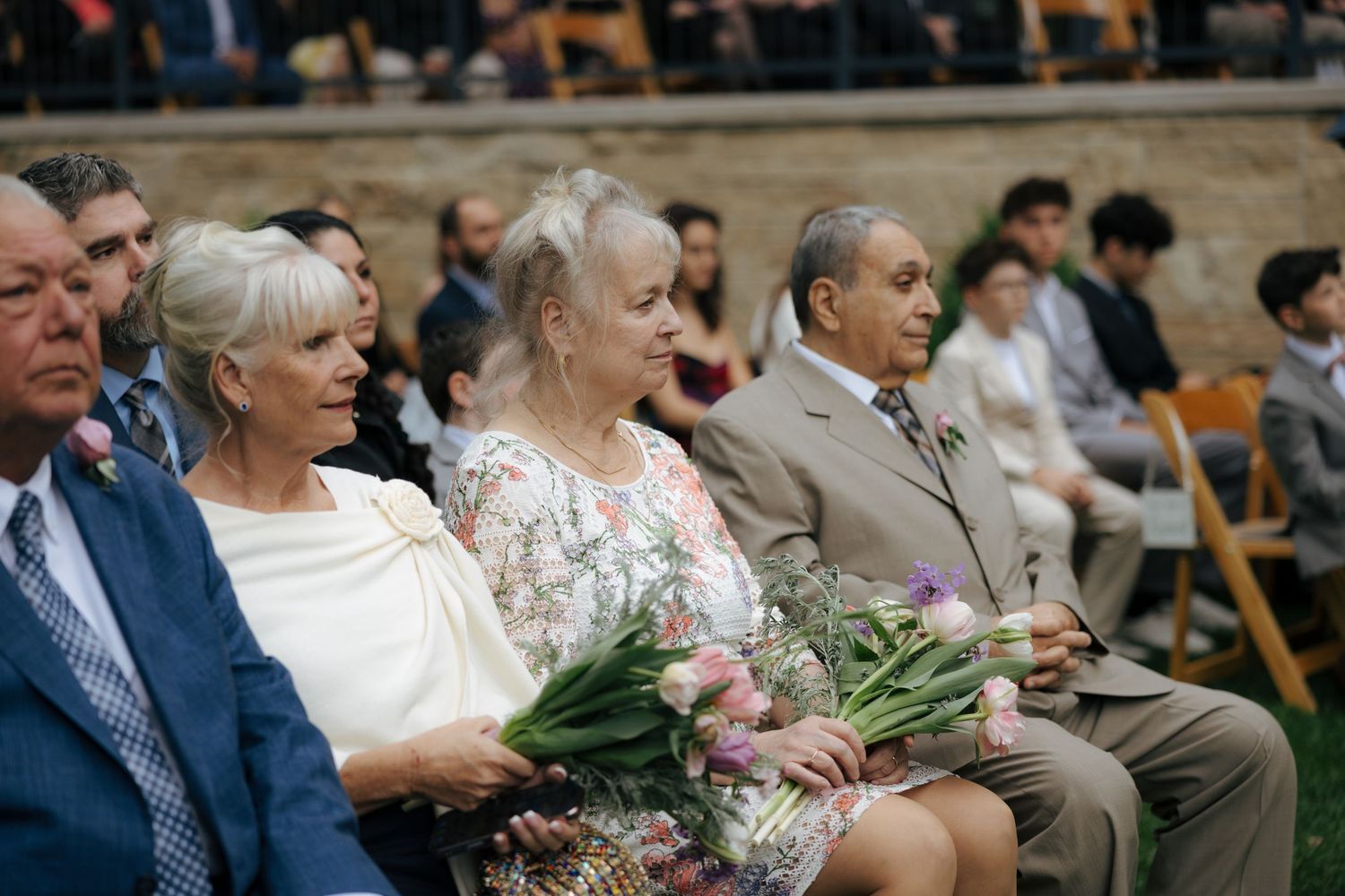 Guests seated at outdoor wedding ceremony, two women holding bouquets of pink tulips, dressed in elegant attire.