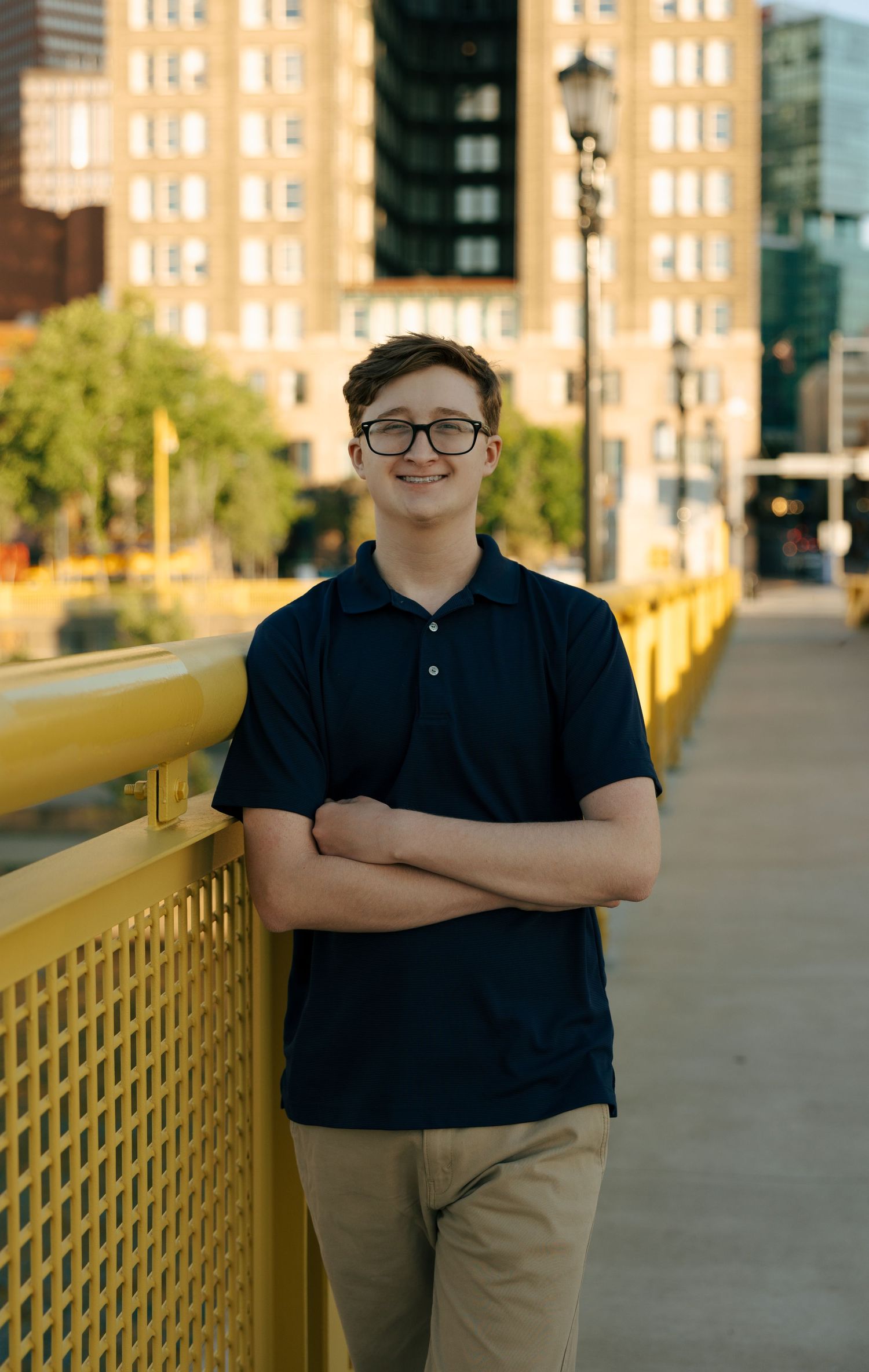 Smiling teen boy with glasses and crossed arms posing on yellow Pittsburgh bridge at golden hour.