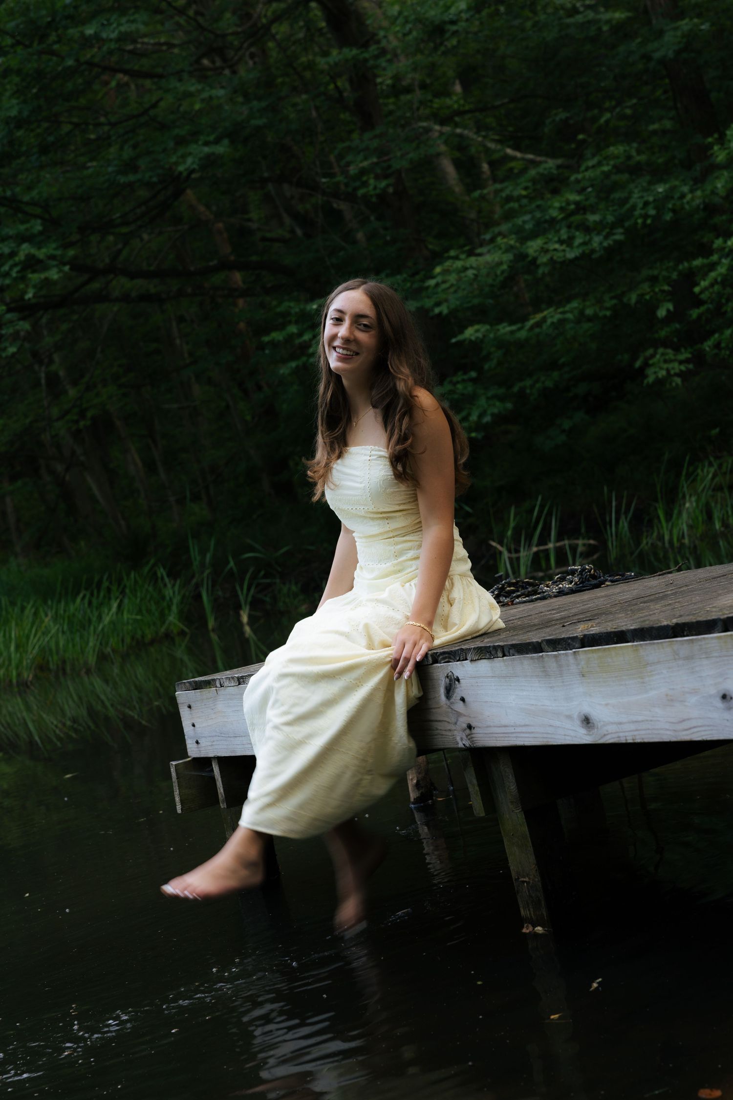 Smiling young woman in cream strapless gown sitting on wooden dock with feet dipping into lake water.
