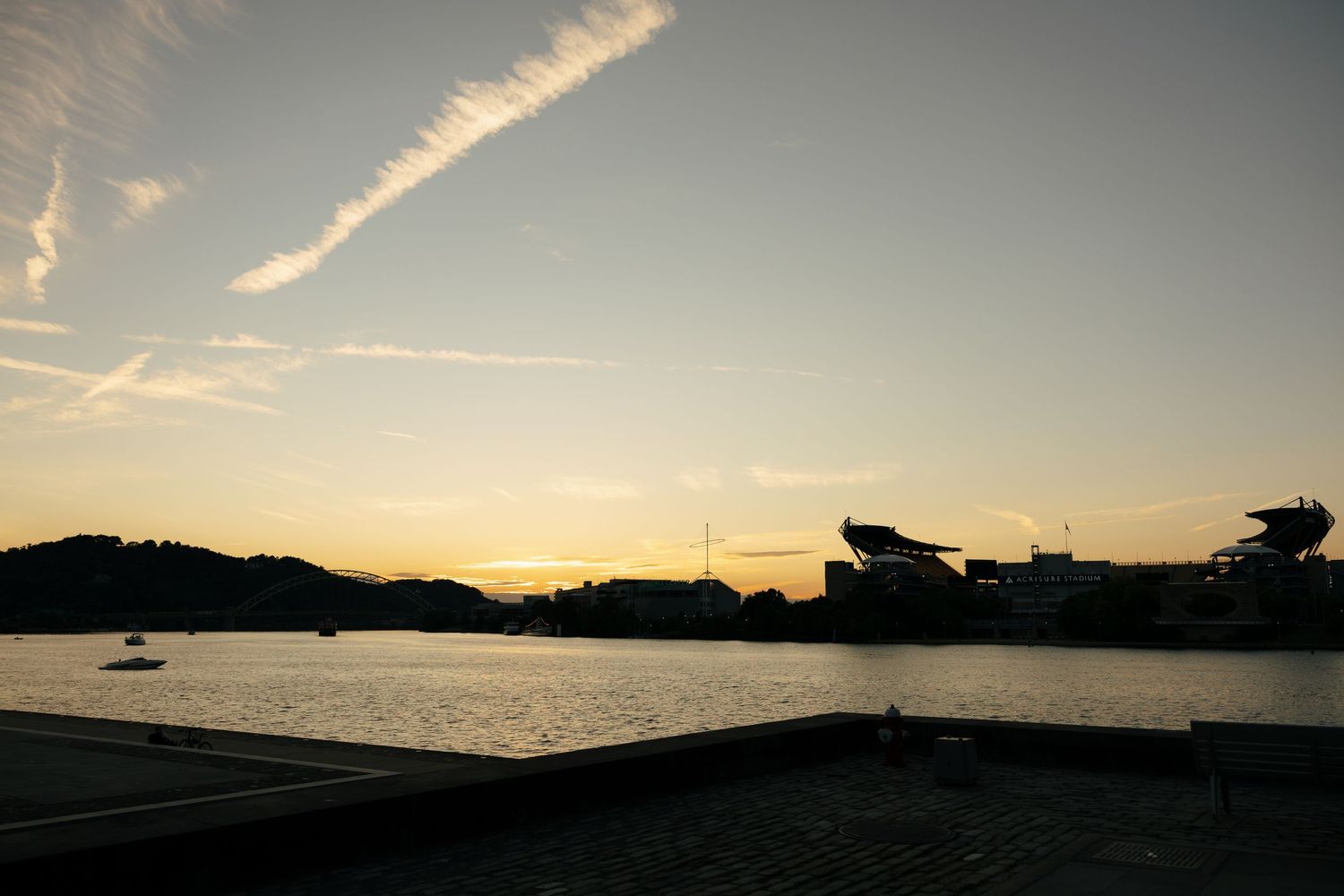 Scenic sunset silhouette of Acrisure Stadium across a calm river with golden sky reflections.