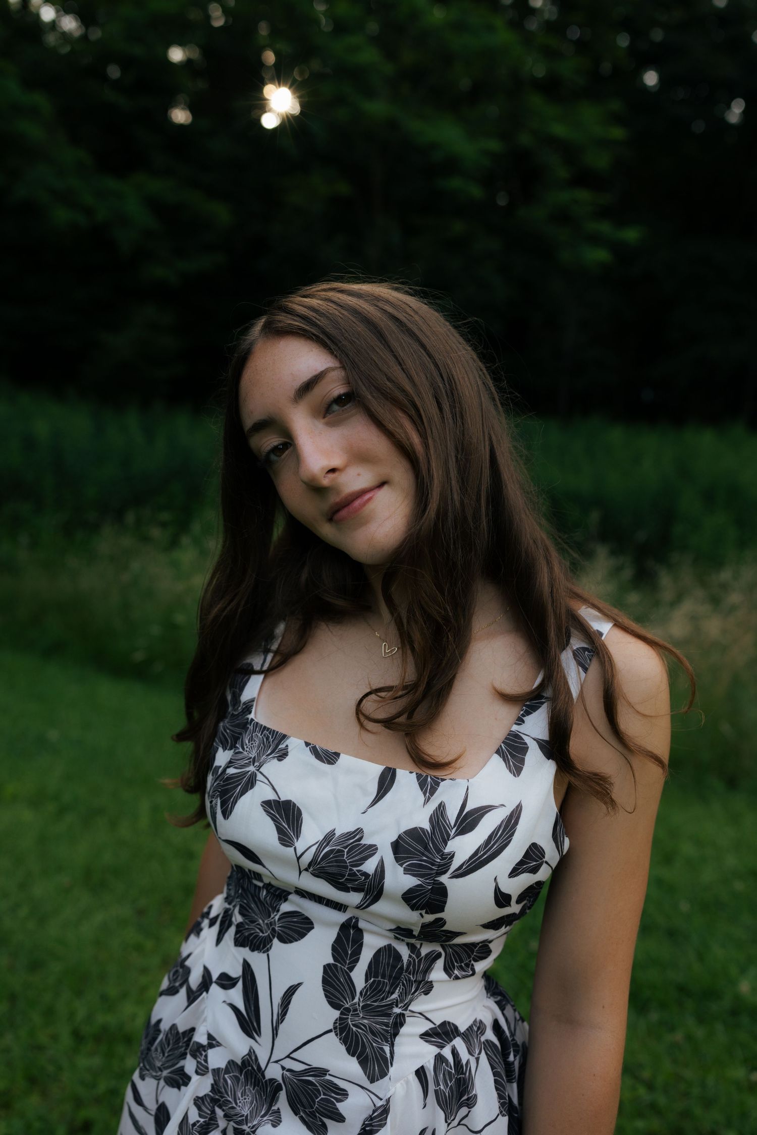Young woman in floral sundress smiling softly outdoors in a green park at twilight.