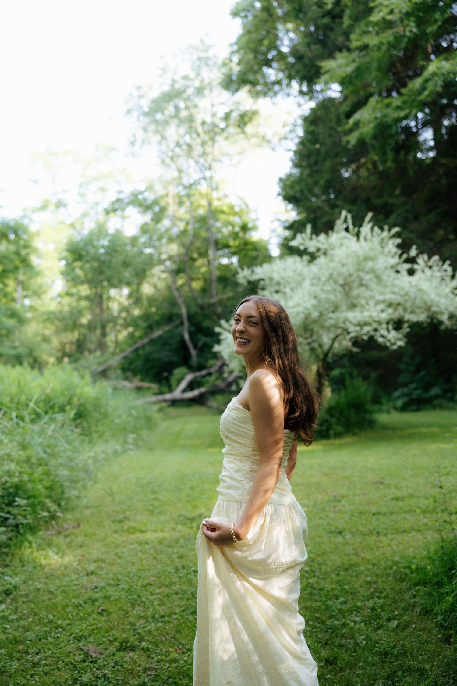 Smiling young woman in elegant cream dress standing in lush green garden, laughing joyfully.