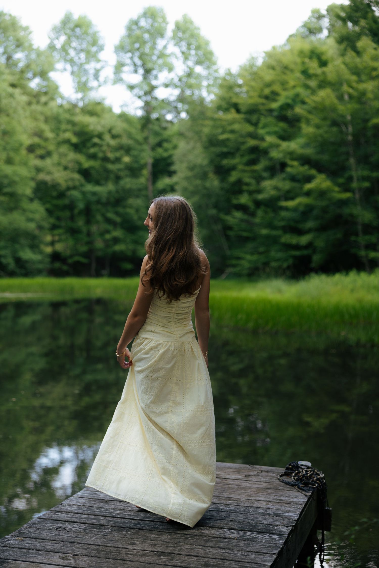 Young woman in cream strapless gown standing on wooden dock overlooking calm reflective lake in forest.