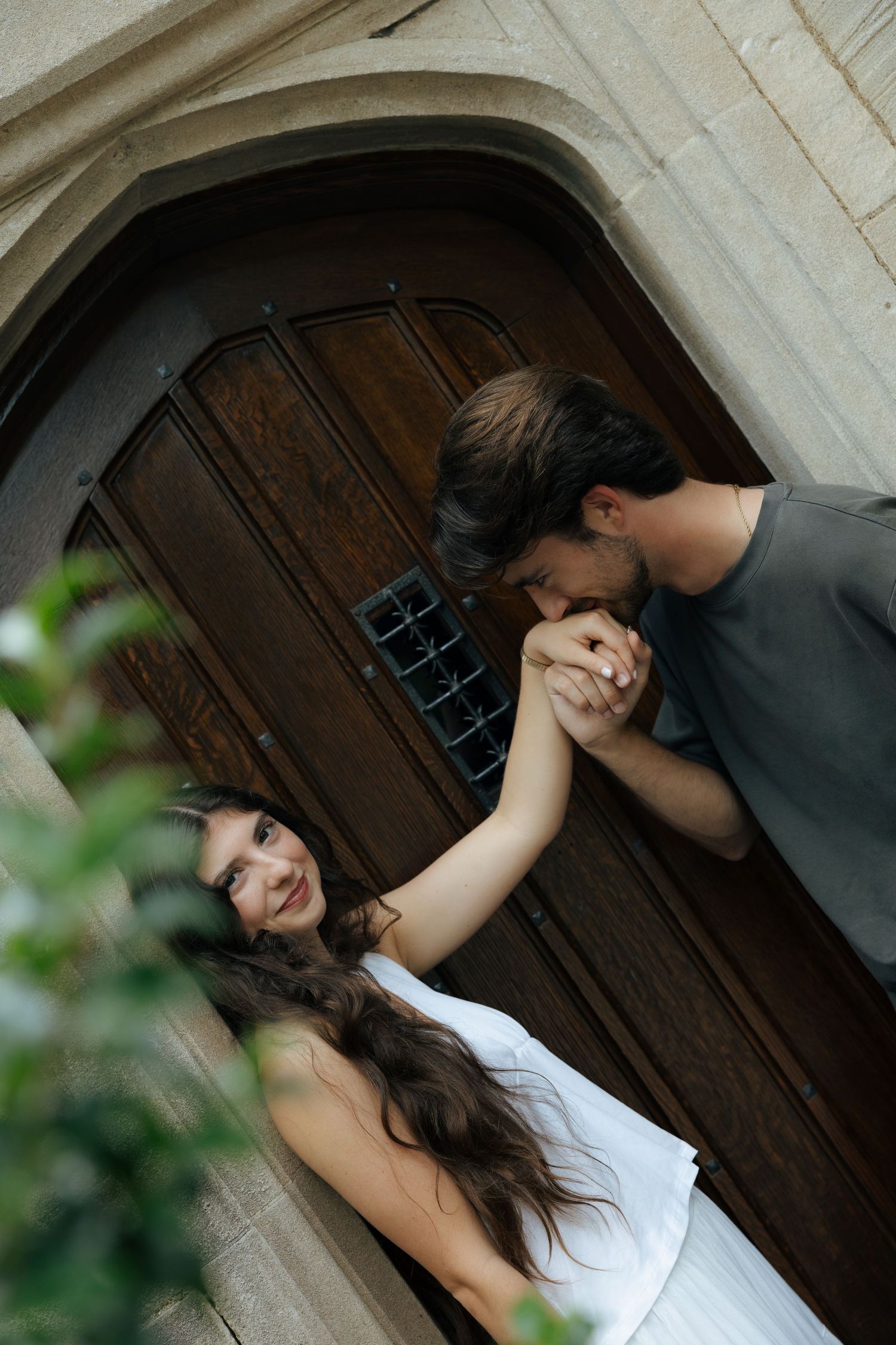 Man kissing woman's hand by ornate wooden arched door in romantic outdoor engagement photo session.