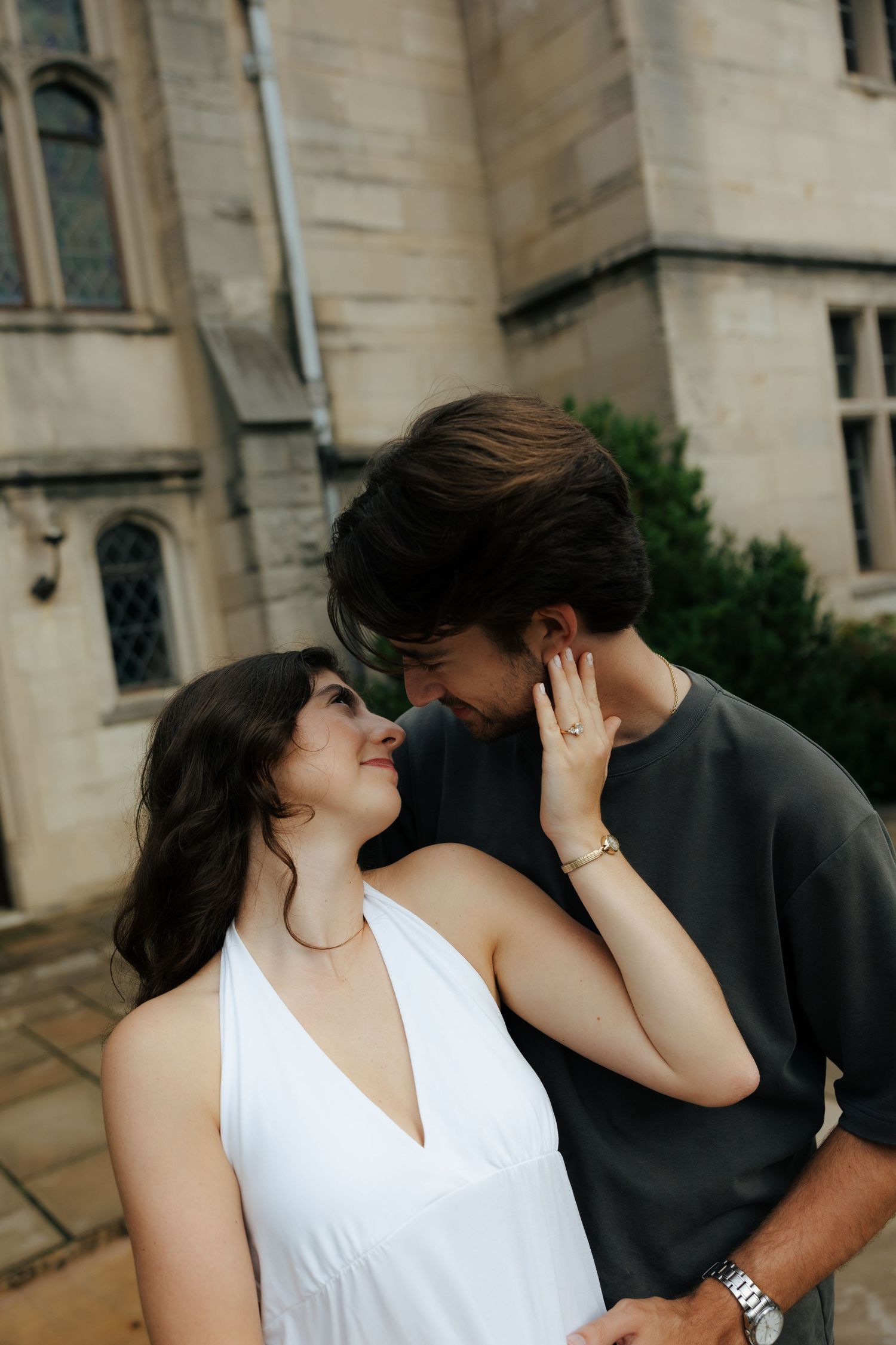 A couple shares a tender, close moment outside a historic stone building, the woman in a white halter dress.
