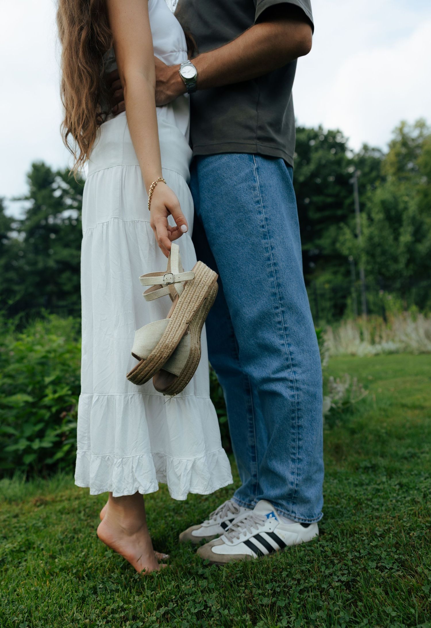 Close-up of couple standing together, woman barefoot holding espadrille sandals in green garden.