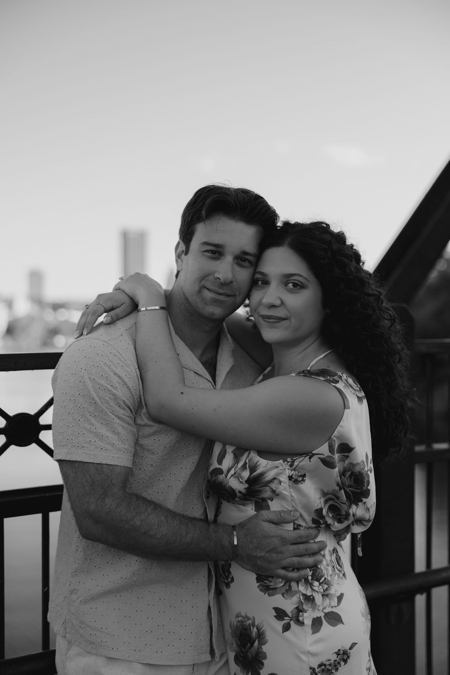 Black and white photo of a couple embracing and smiling near a waterfront railing.