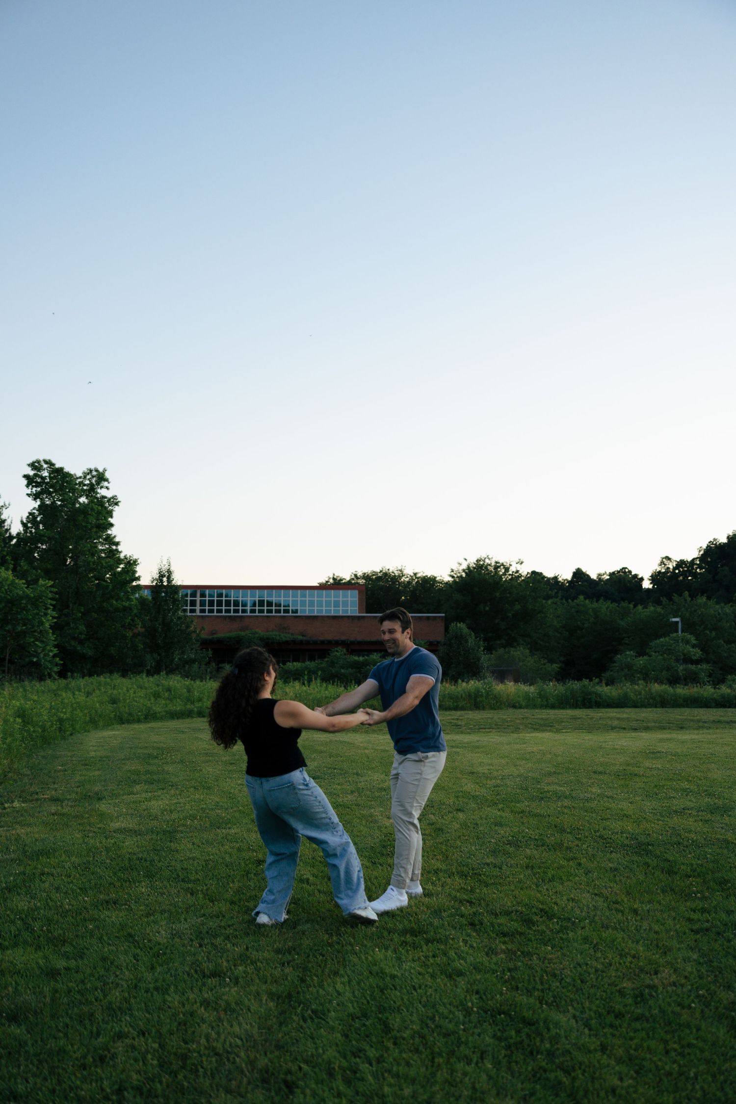 Couple dancing and spinning together on a green lawn at dusk with trees and a brick building in the background.