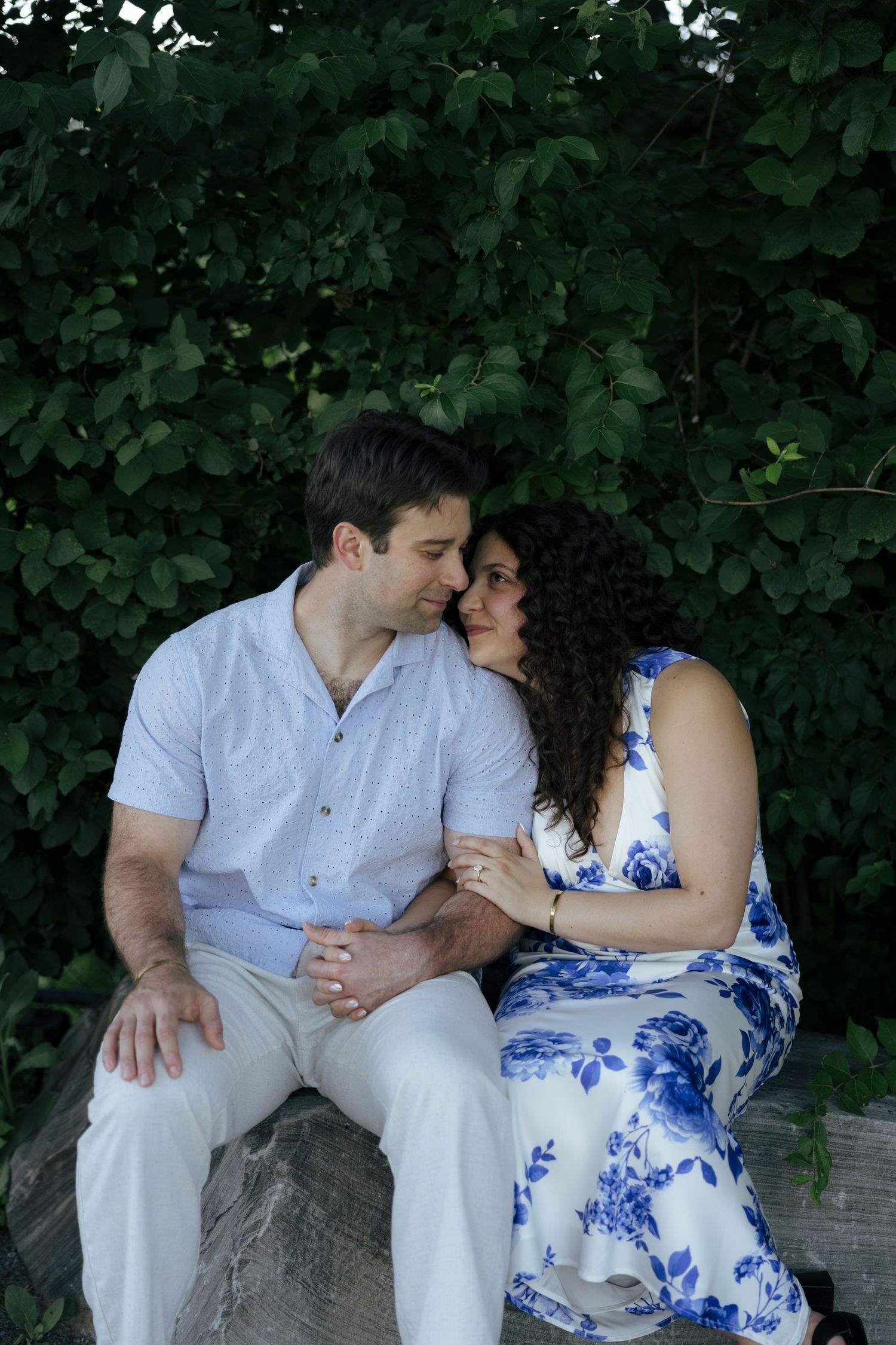 Couple sitting close together on stone bench surrounded by lush green foliage.